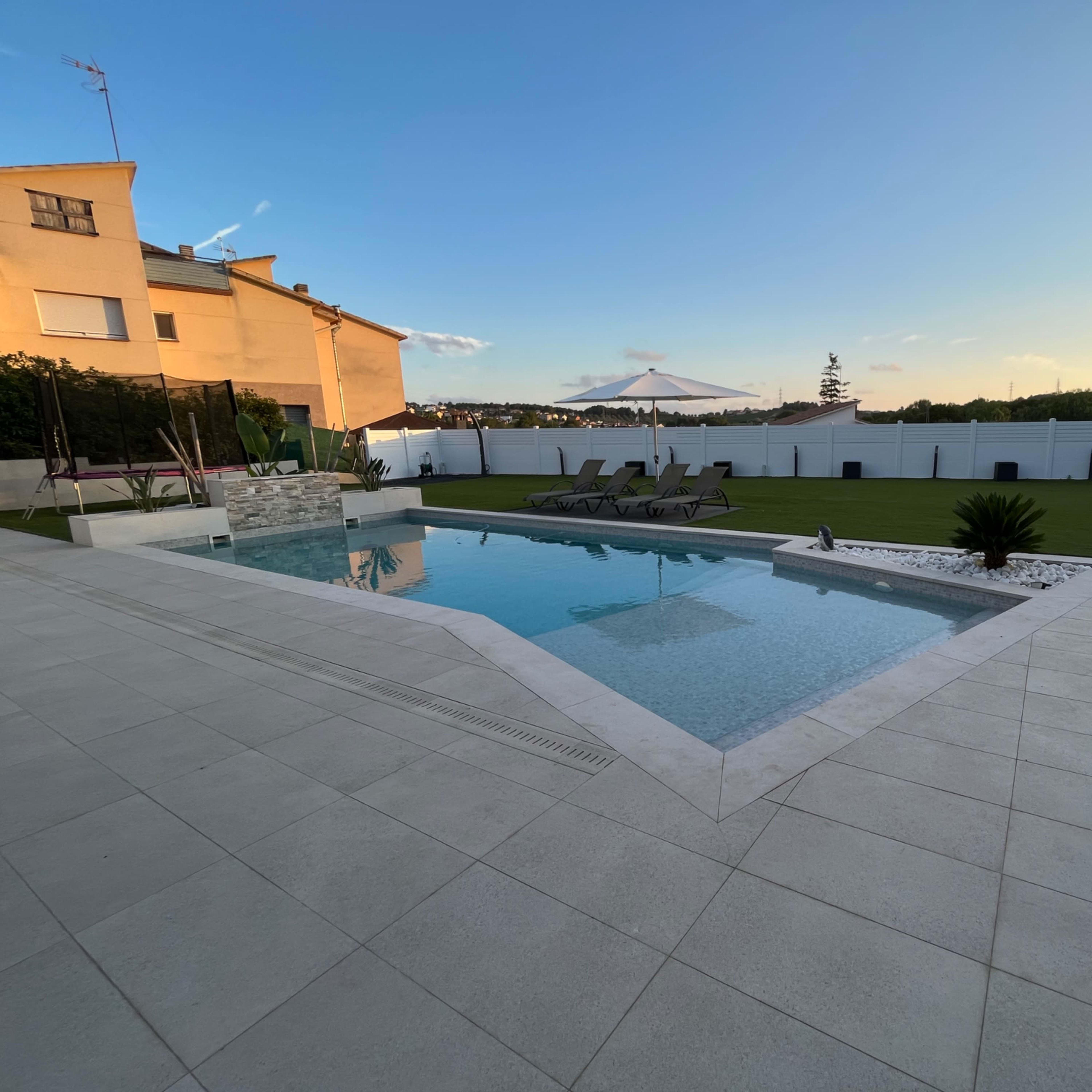 A modern backyard with a swimming pool surrounded by a paved area, loungers, and a grass lawn.