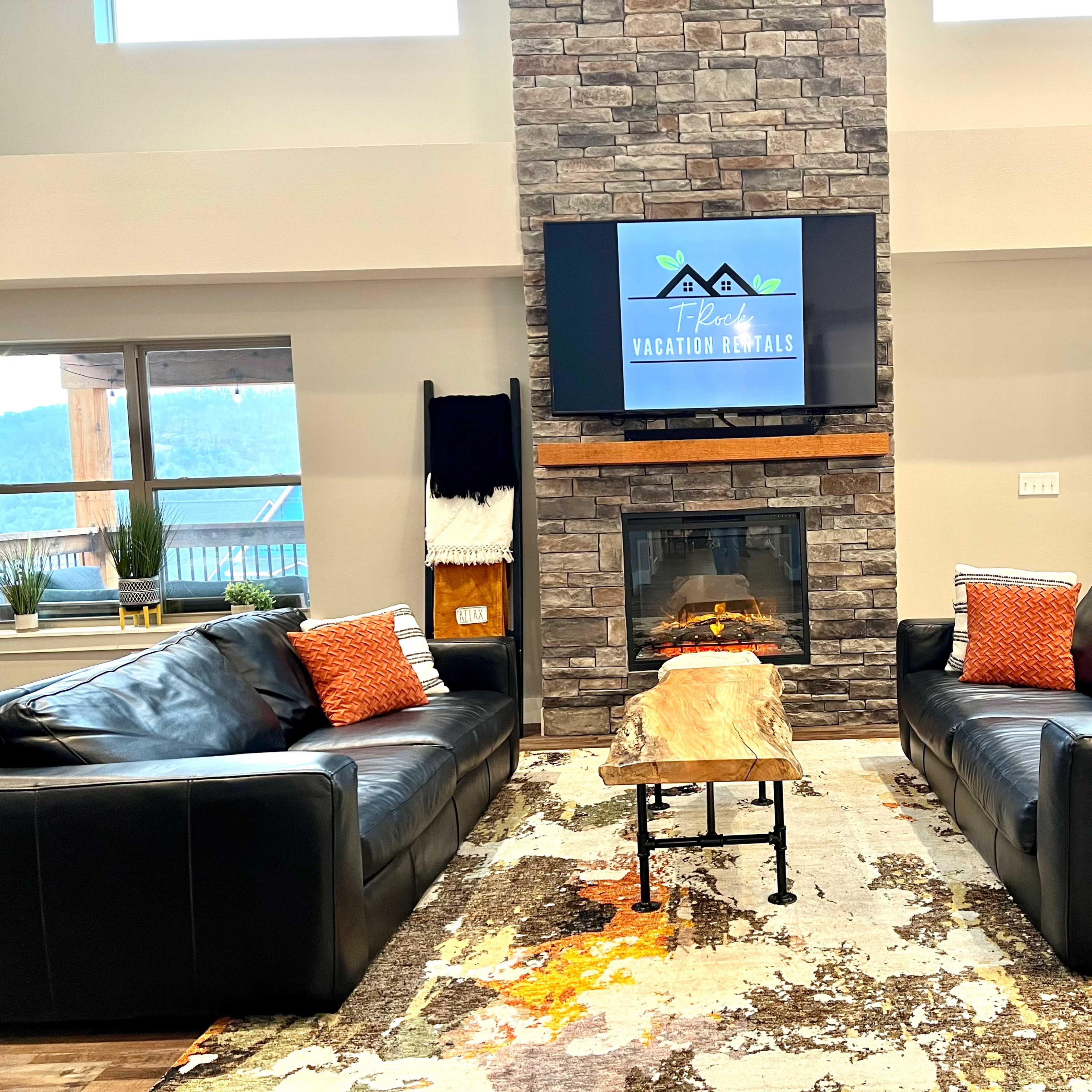 A living room with two black leather sofas facing a stone fireplace beneath a wall-mounted television, surrounded by a patterned rug and large windows letting in natural light.