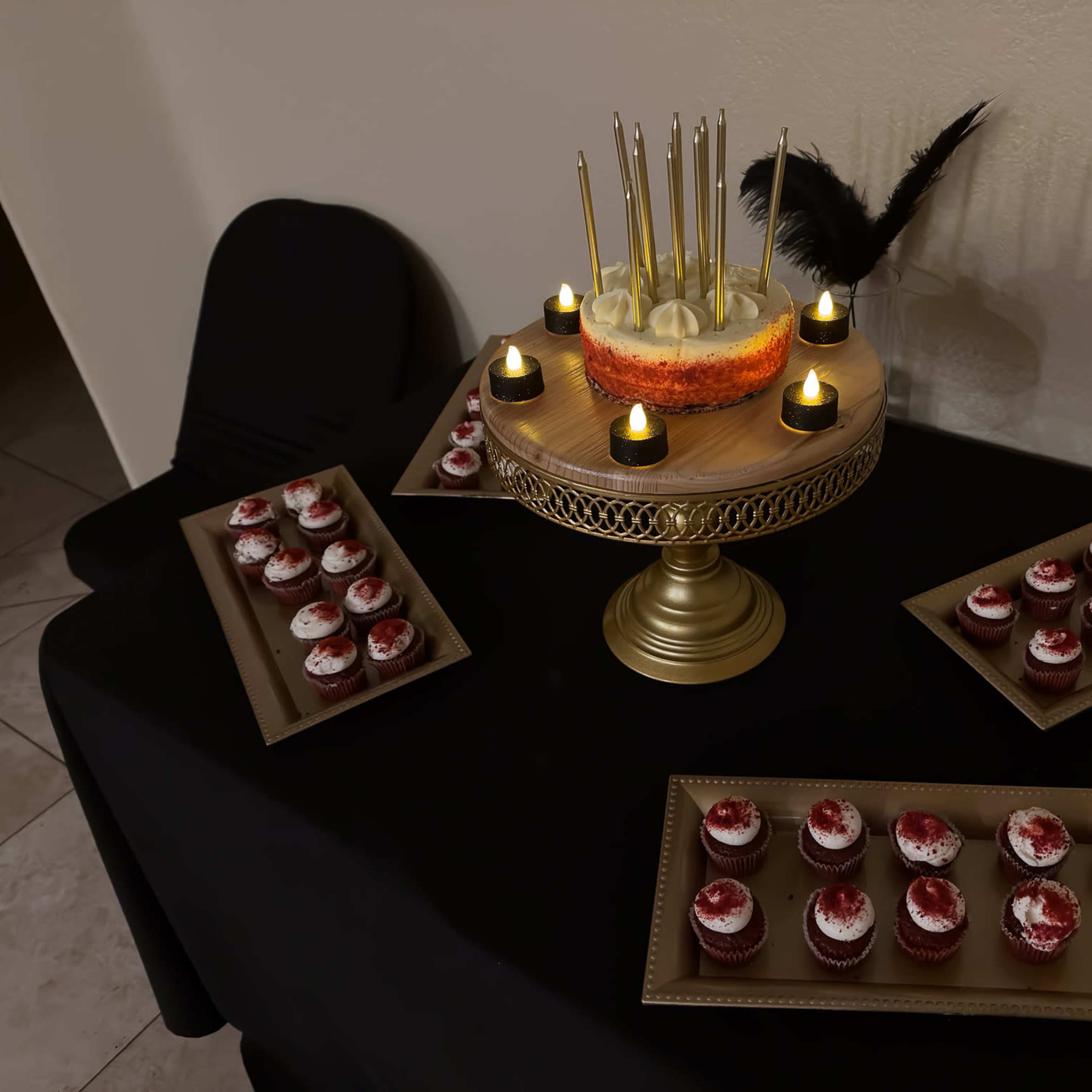 A table covered with a black cloth displays a tiered cake adorned with lit candles and several trays of red velvet cupcakes.