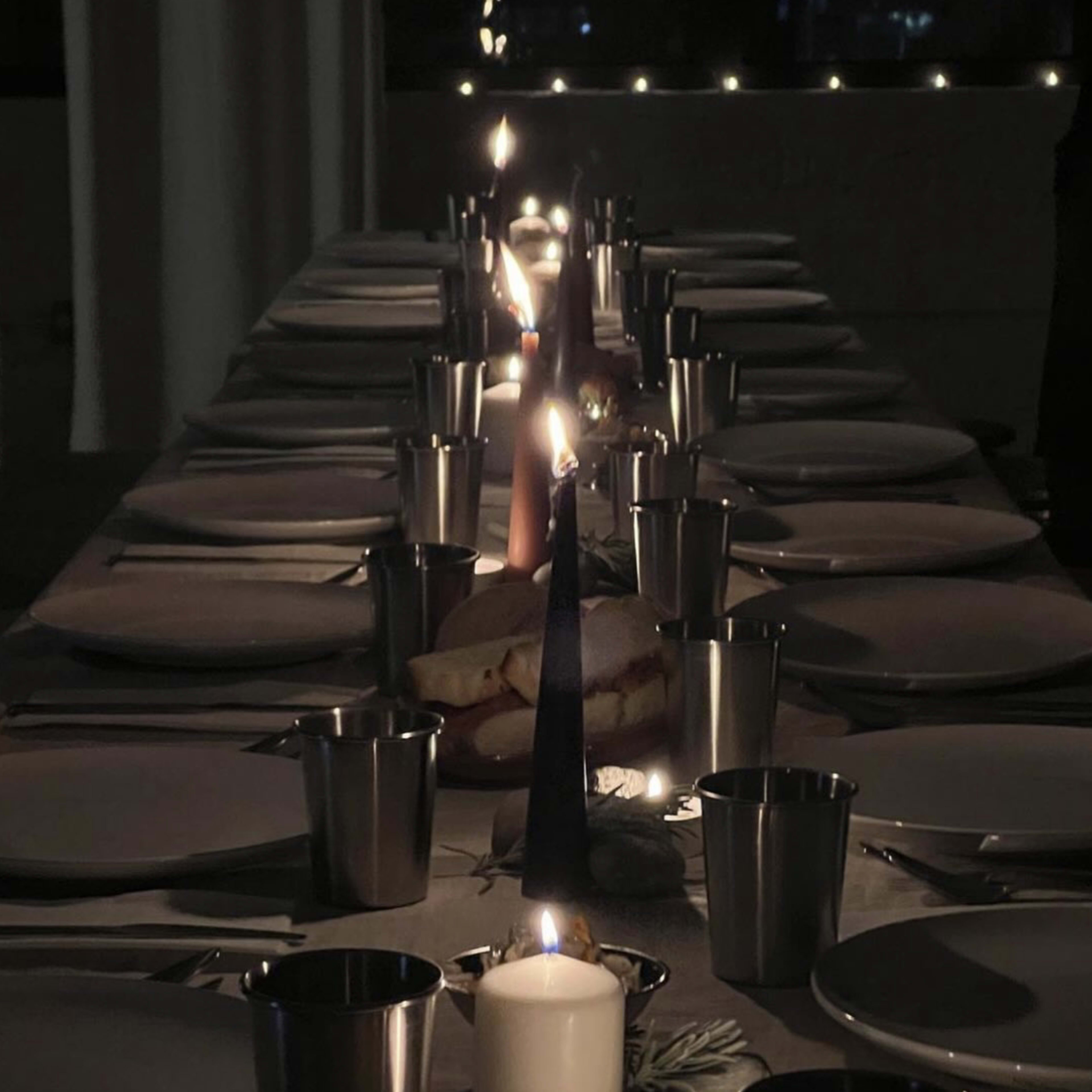 A long dining table is set with plates and metal cups, illuminated by several candles.
