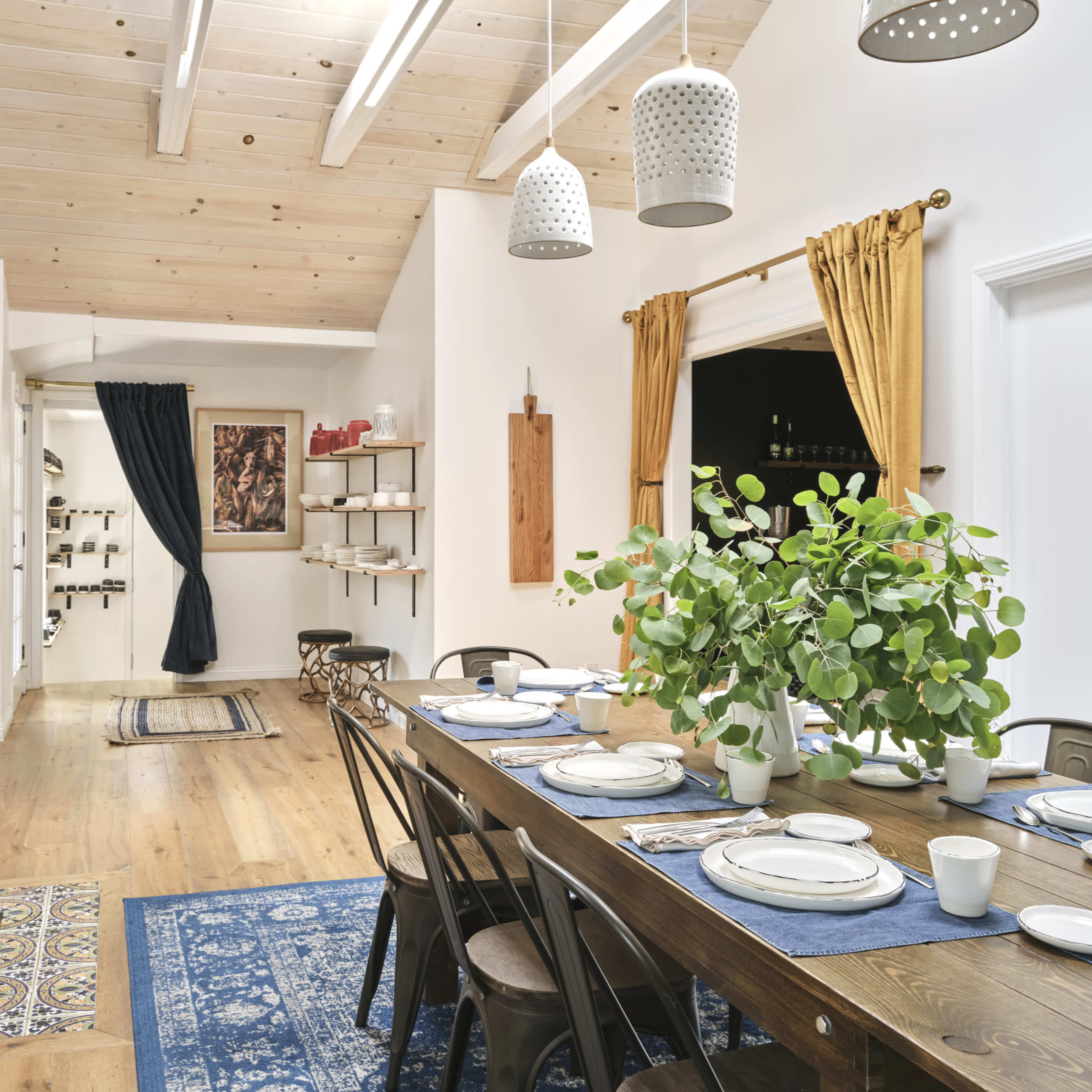 A modern dining area with a long wooden table set for a meal, surrounded by chairs, under pendant lights and a wooden ceiling.