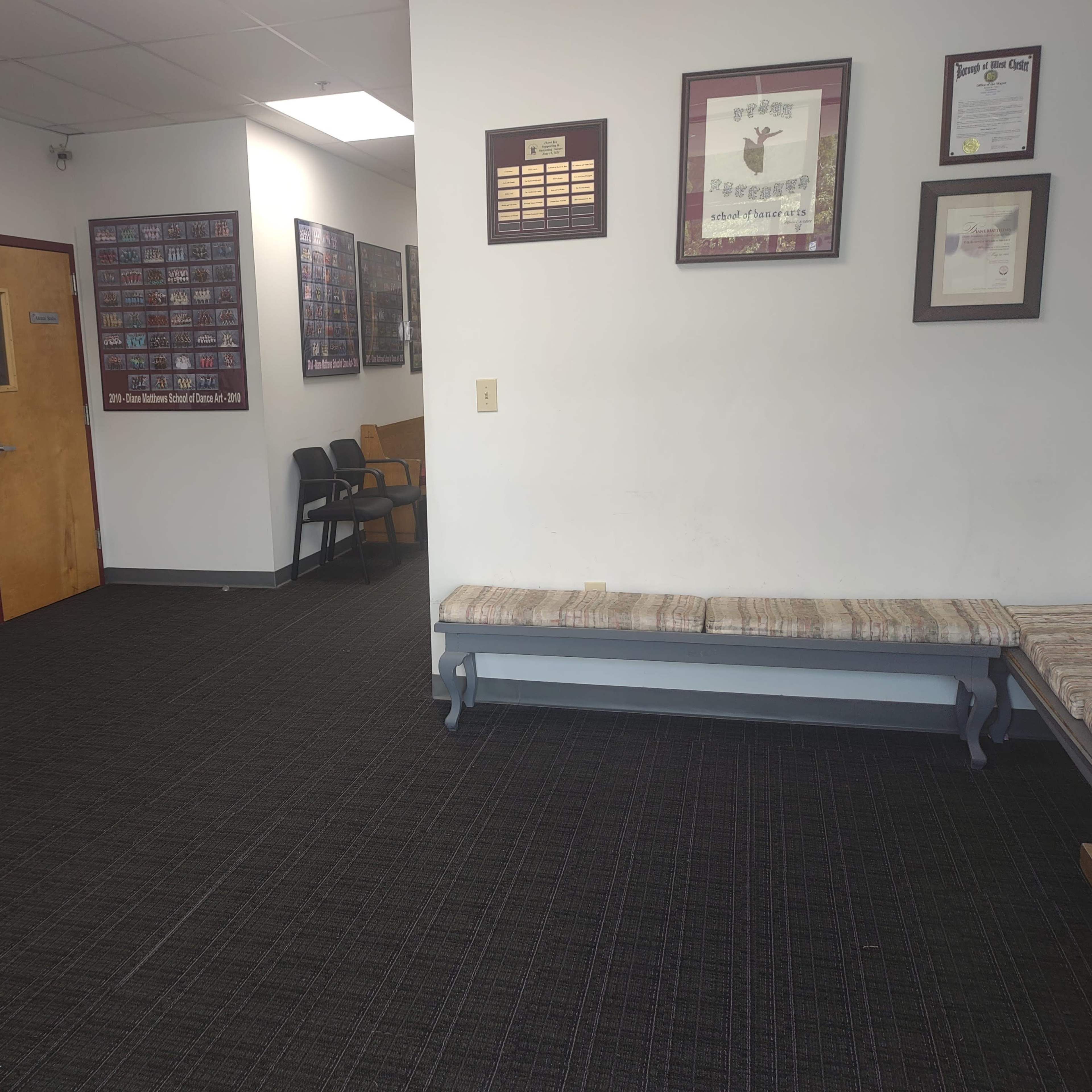 A waiting area with a wall-mounted display of plaques and framed photographs, a long bench against one wall, and a few chairs arranged nearby.