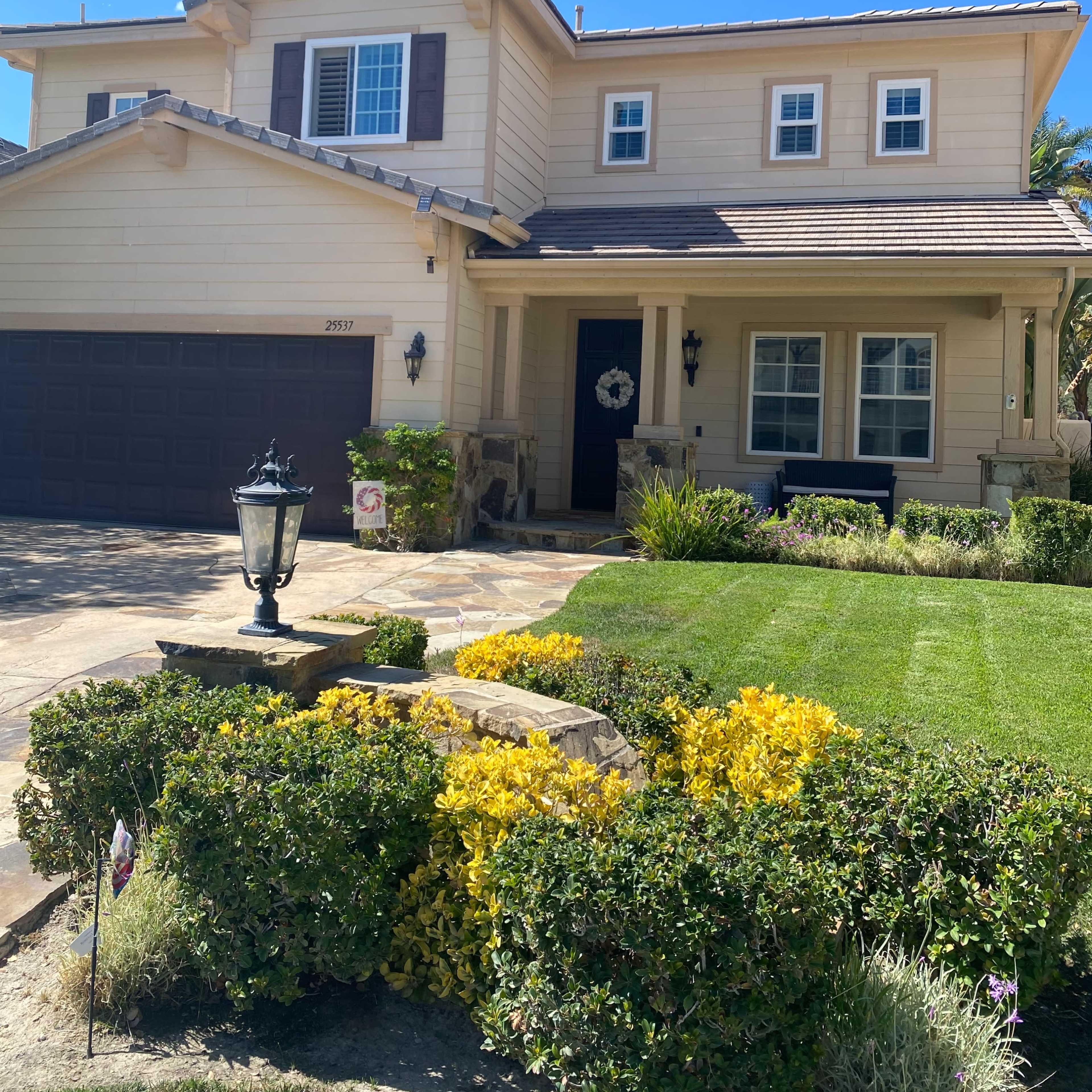 A two-story beige house with dark shutters and a landscaped front yard features a garage and a porch with decorative elements.