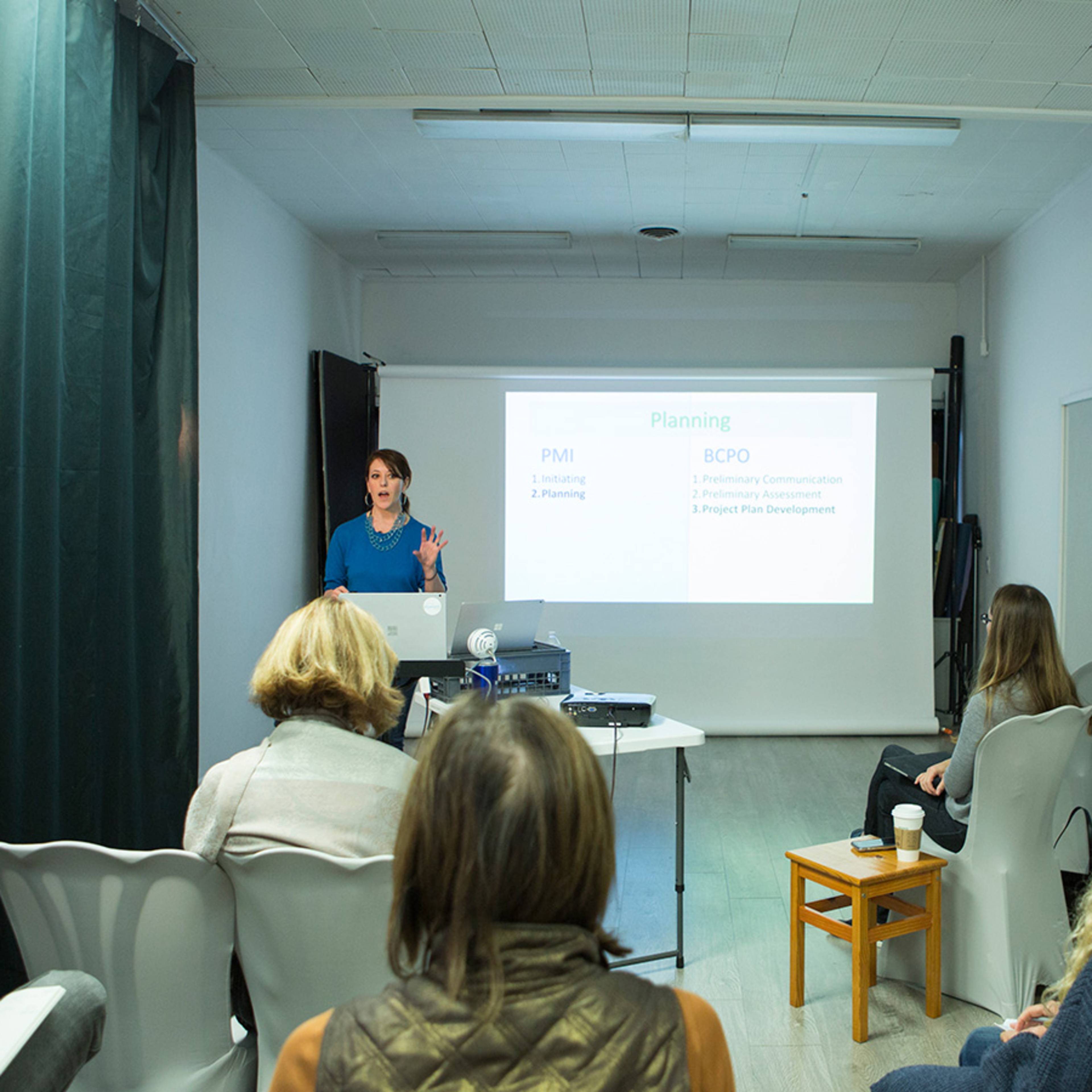 A woman delivers a presentation in a bright room, facing an audience seated in chairs, with a projector displaying slides on a screen.