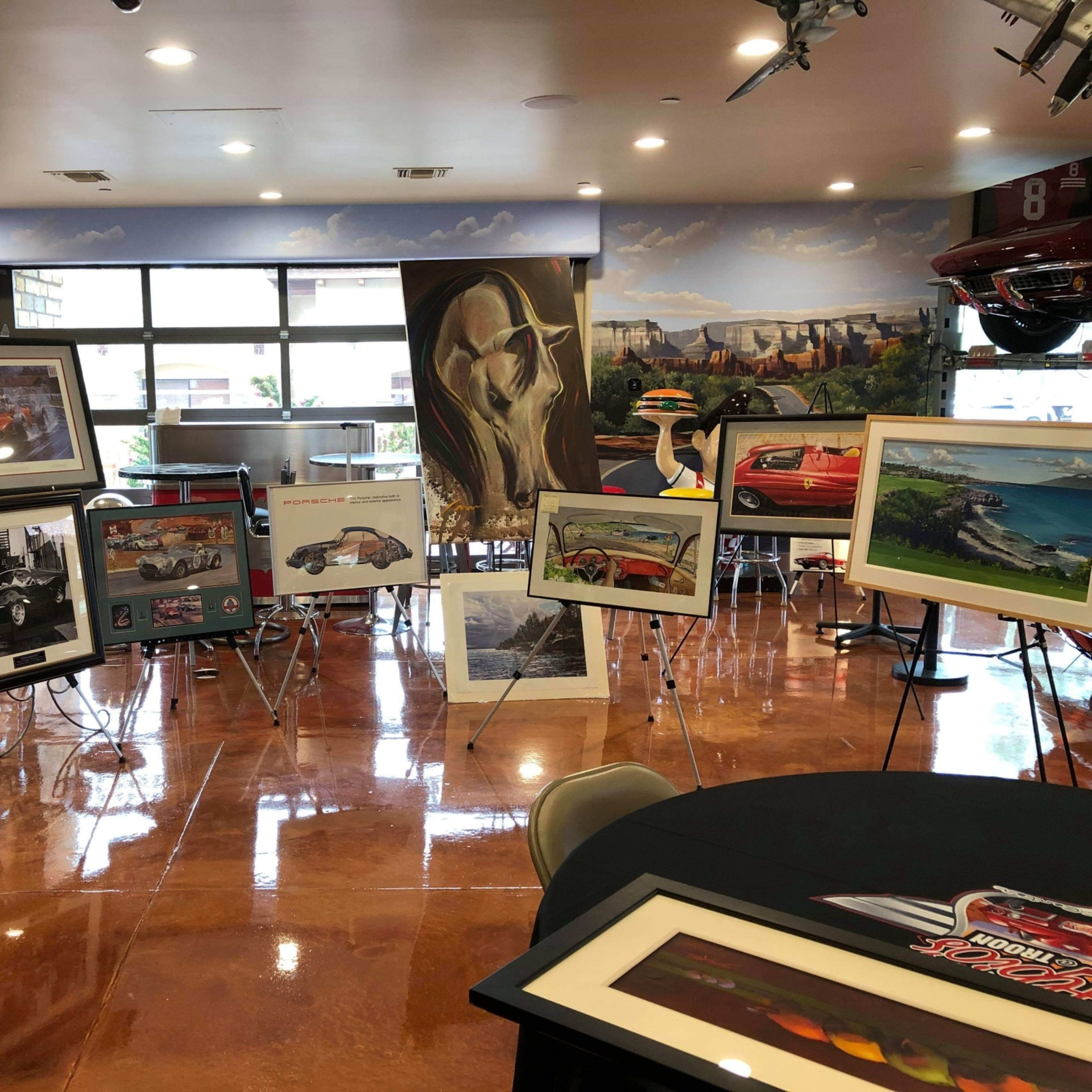 An interior space with various framed artworks displayed on easels, alongside a round table and vintage car memorabilia.