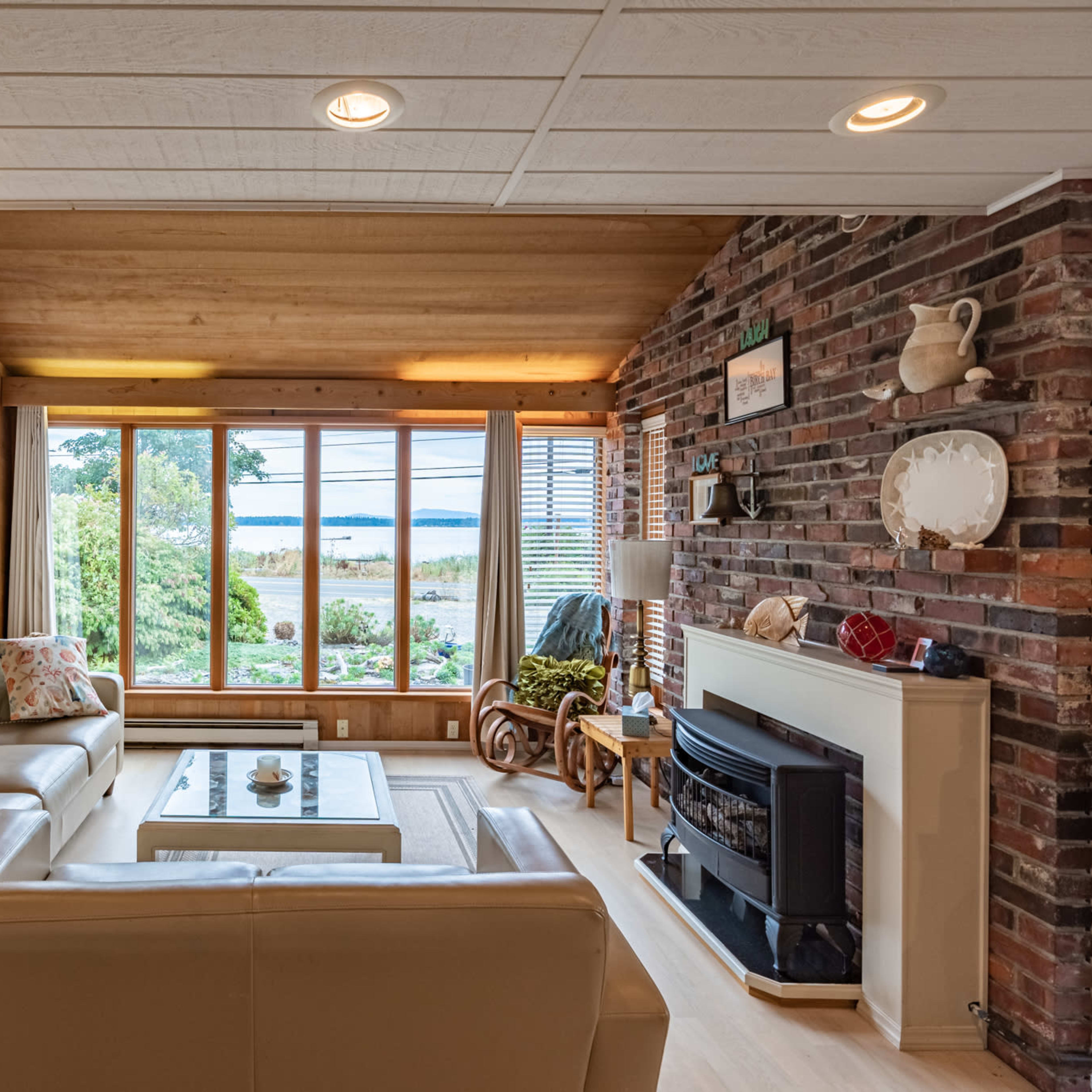 The living room features a beige sofa set, a lamp, and a fireplace, with large windows offering a view of the garden and water beyond.