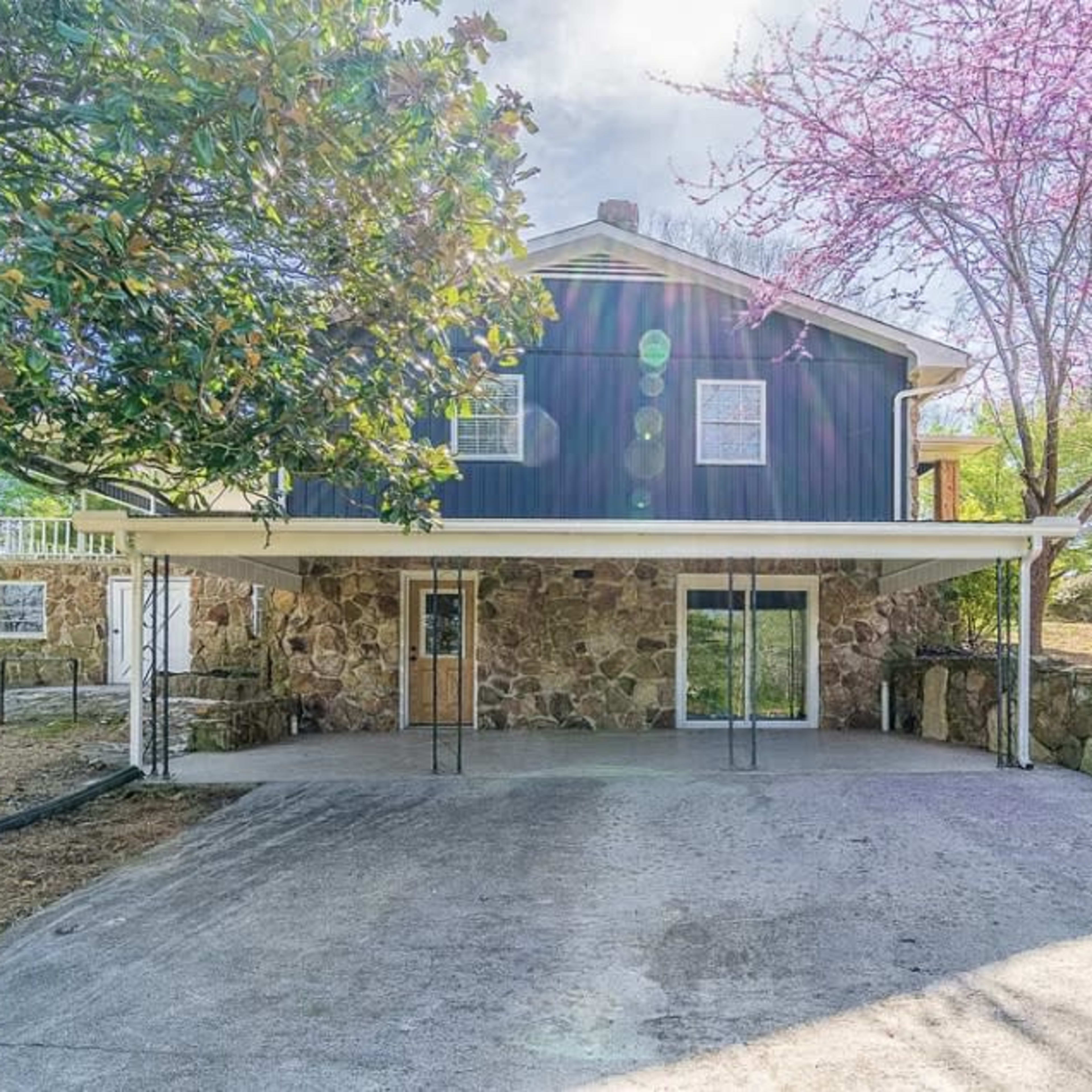 A two-story house with a stone facade and a covered patio is situated in a grassy area with blooming trees.