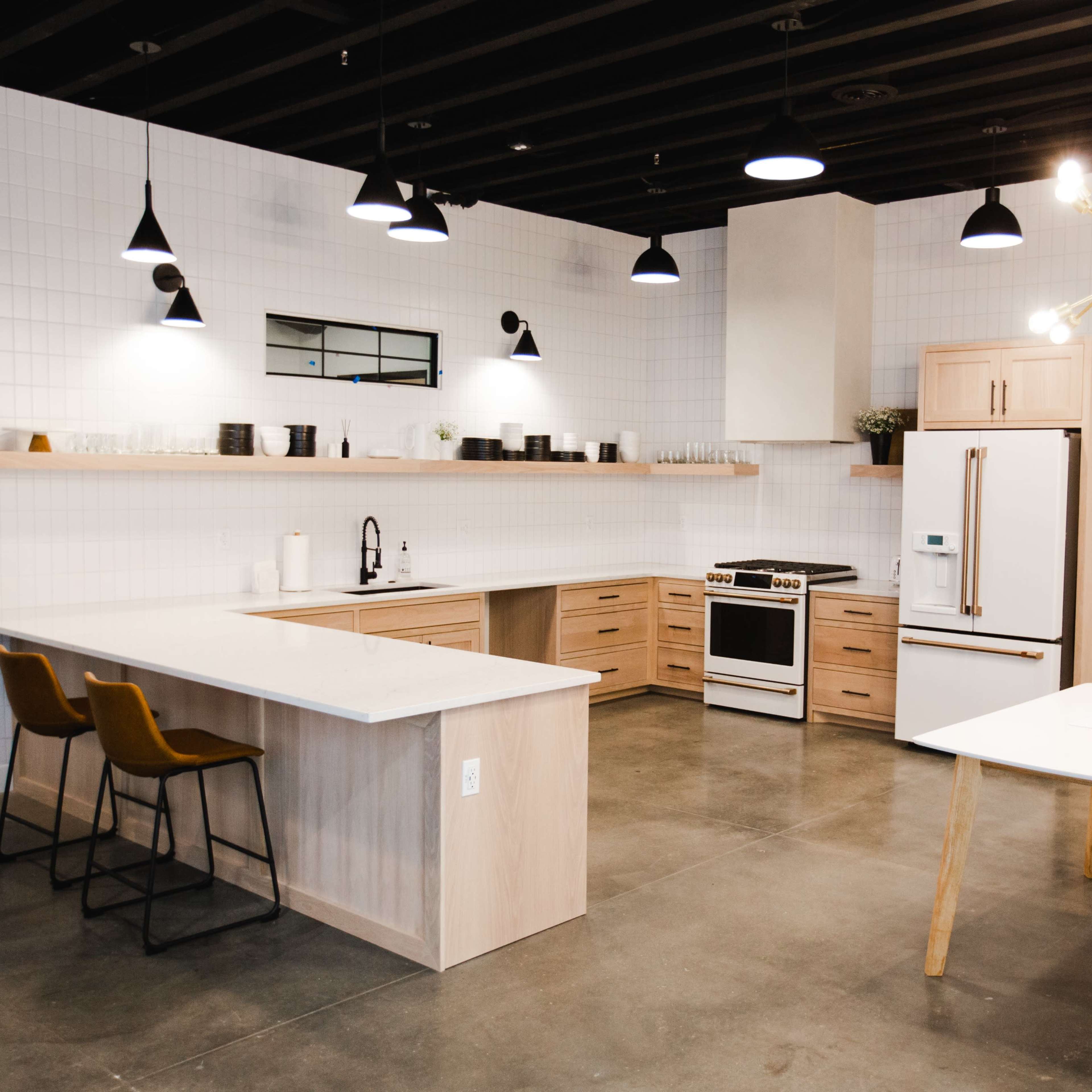 A modern kitchen with a central island, a refrigerator, an oven, and a dining table surrounded by chairs, all set against a backdrop of white tiled walls.
