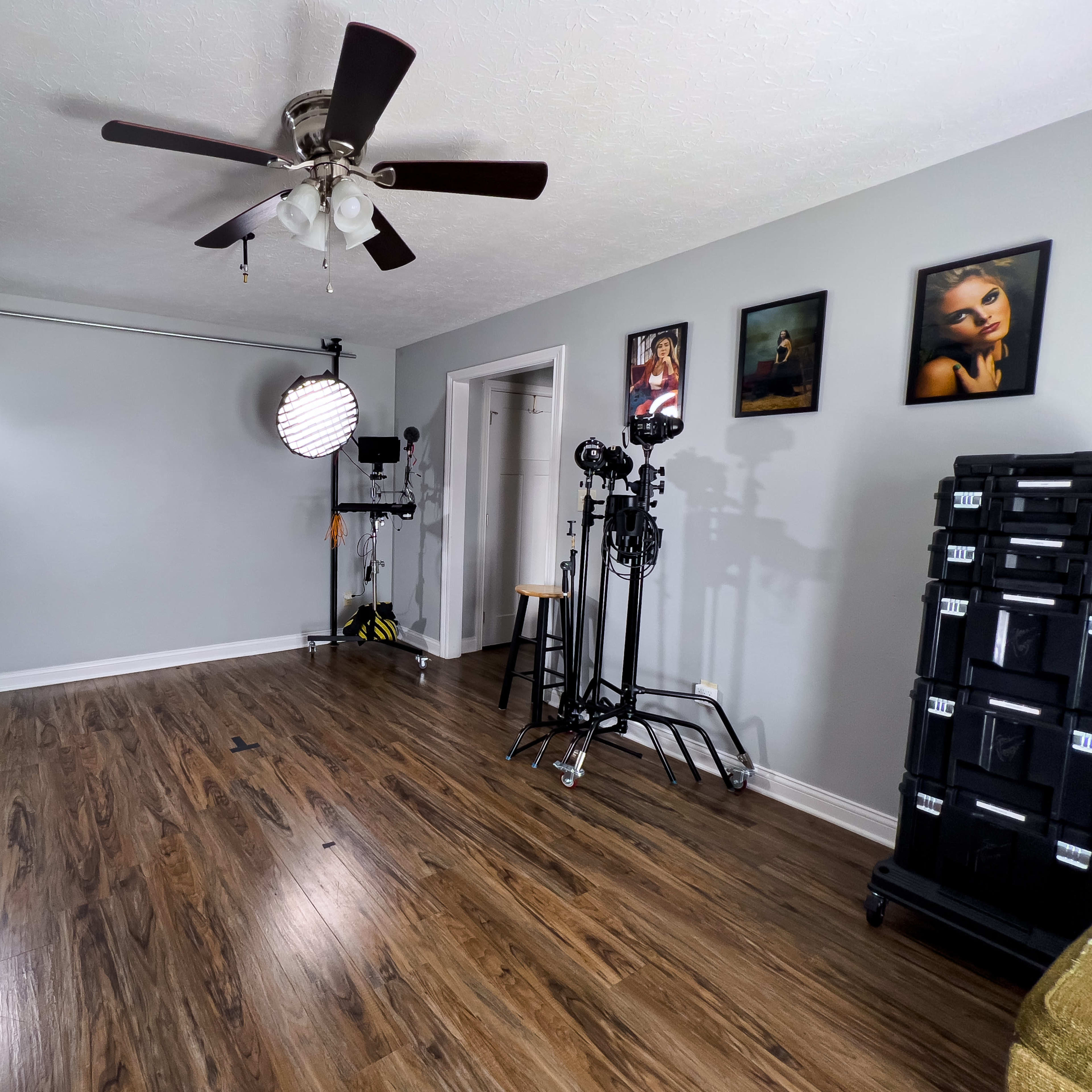 A spacious room with hardwood floors, featuring photography equipment, a ceiling fan, and several framed artworks on the walls.