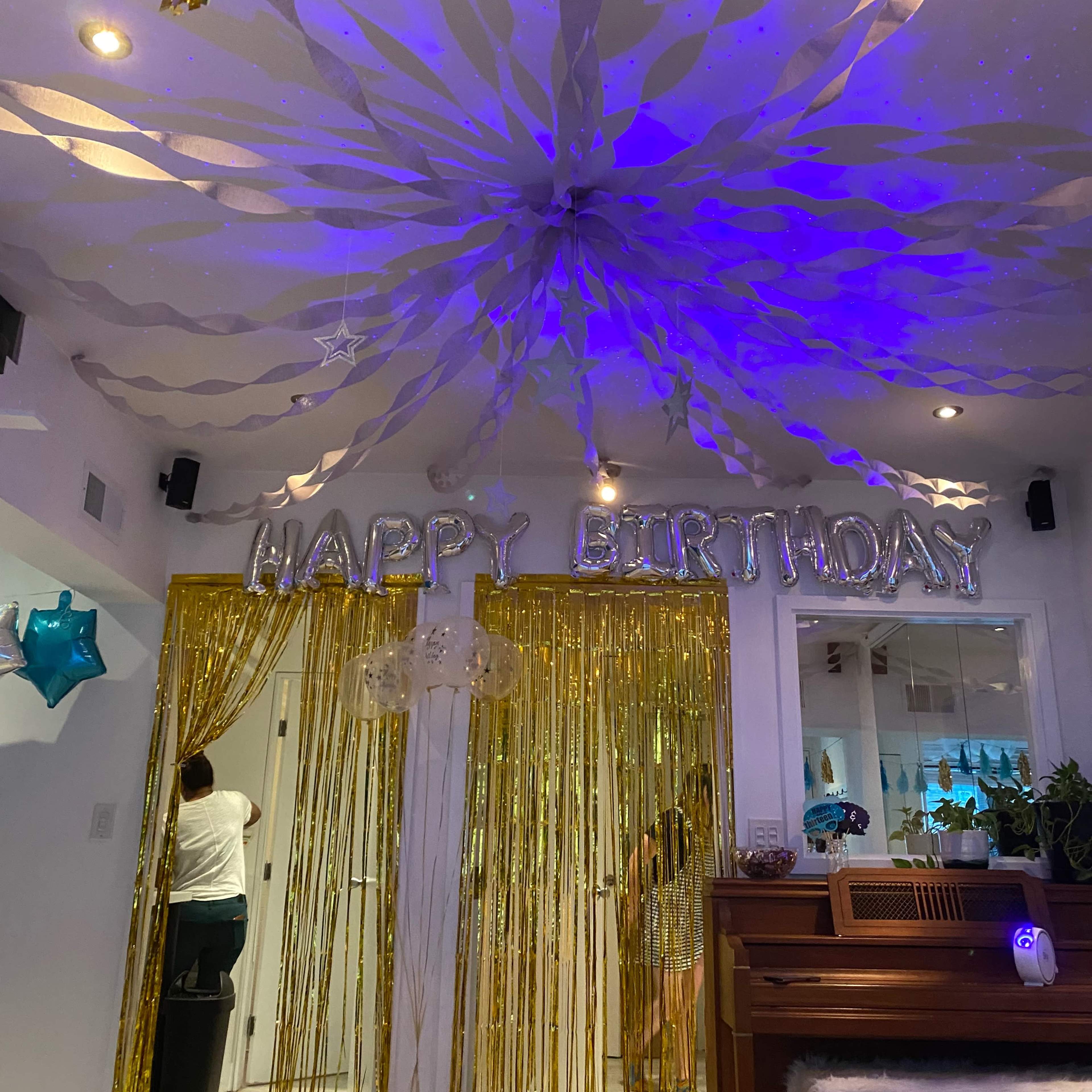 A decorated interior space for a birthday celebration, featuring a "HAPPY BIRTHDAY" banner, metallic curtains, and colorful streamers hanging from the ceiling.