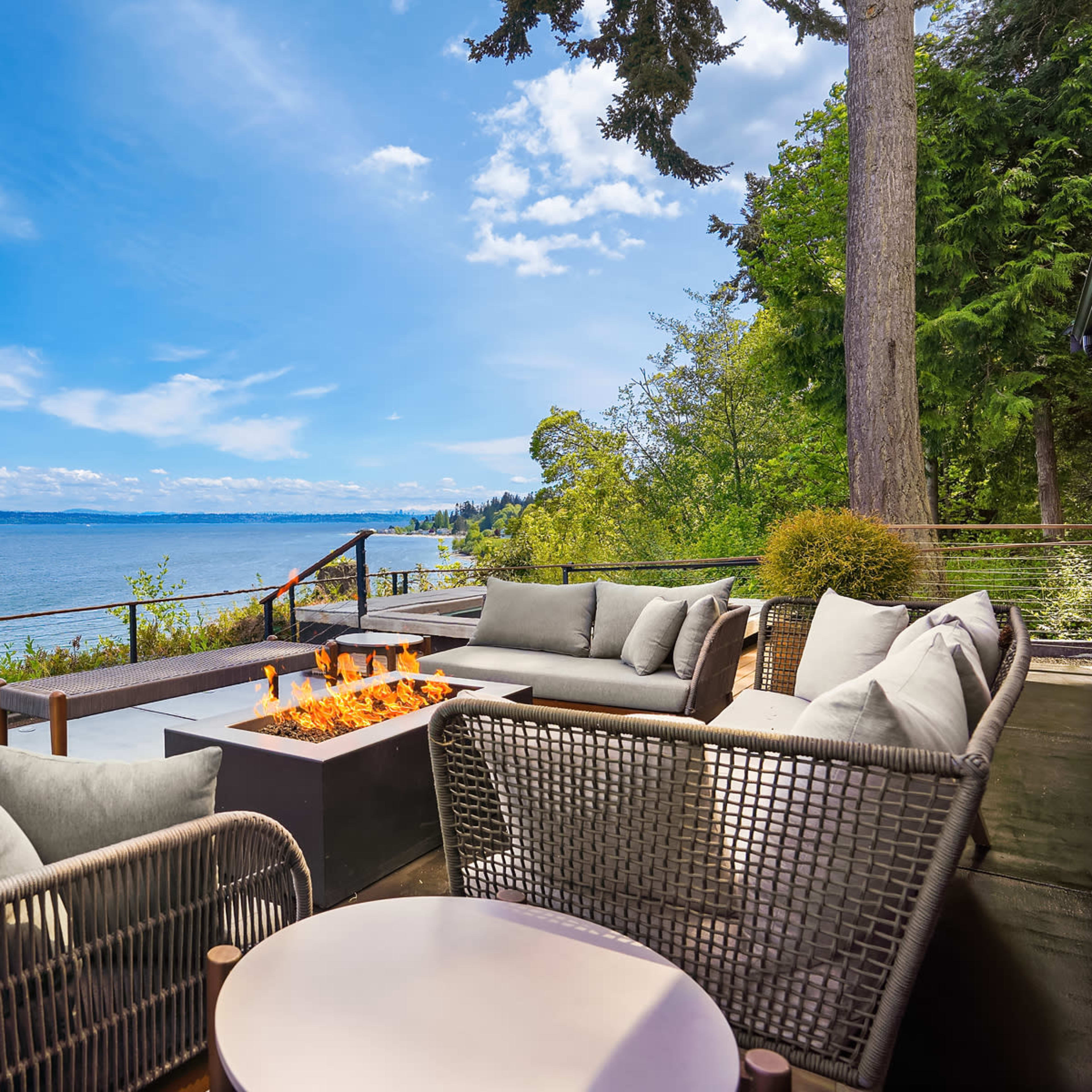 A modern patio with comfortable seating and a fire pit overlooks a calm lake under a blue sky with scattered clouds.