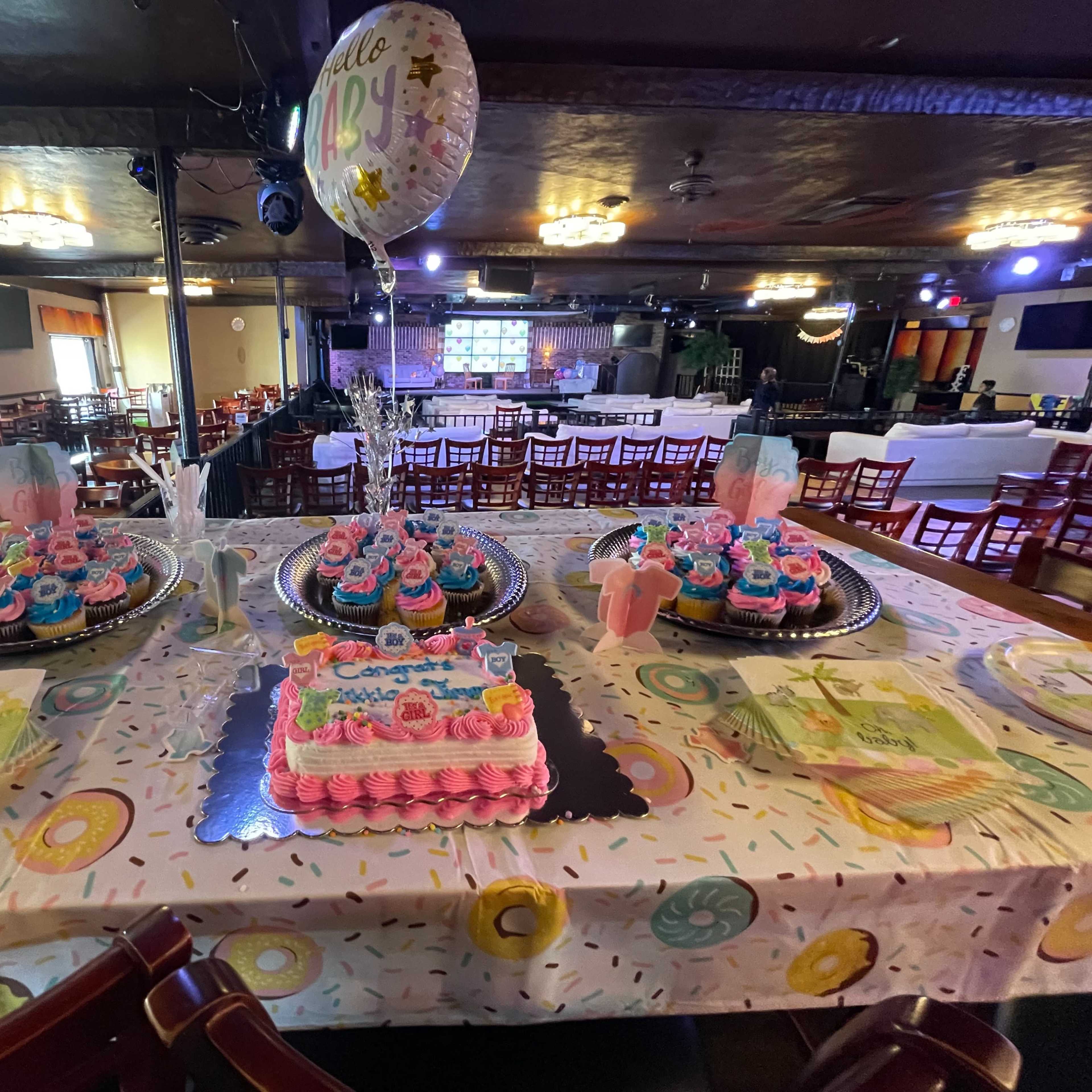 A decorated table set up for a celebration, featuring multiple colorful cakes, cupcakes, and themed tableware in a venue with dim lighting.