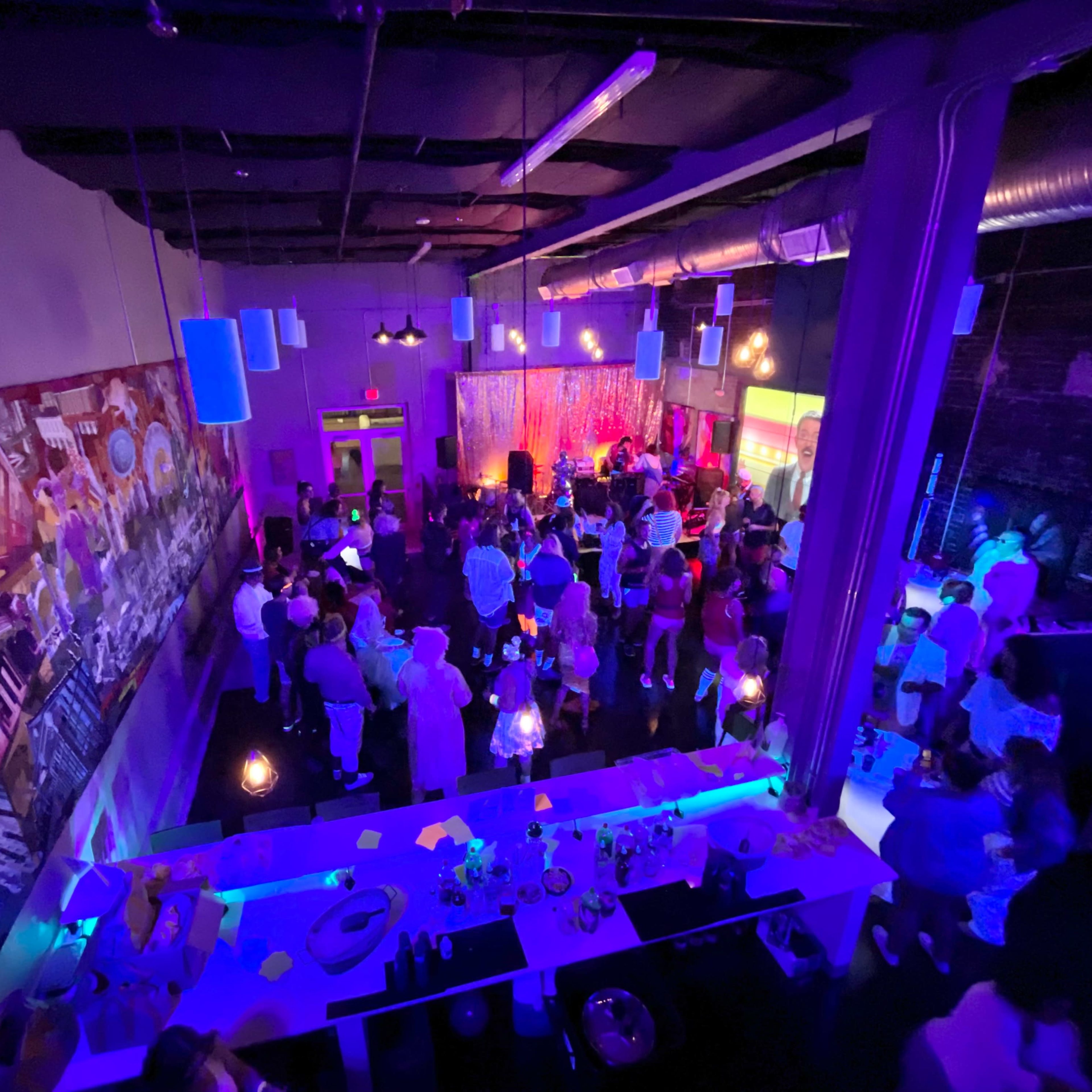 A crowded indoor venue features vibrant lighting and a large mural on the wall, with people socializing and enjoying the atmosphere.