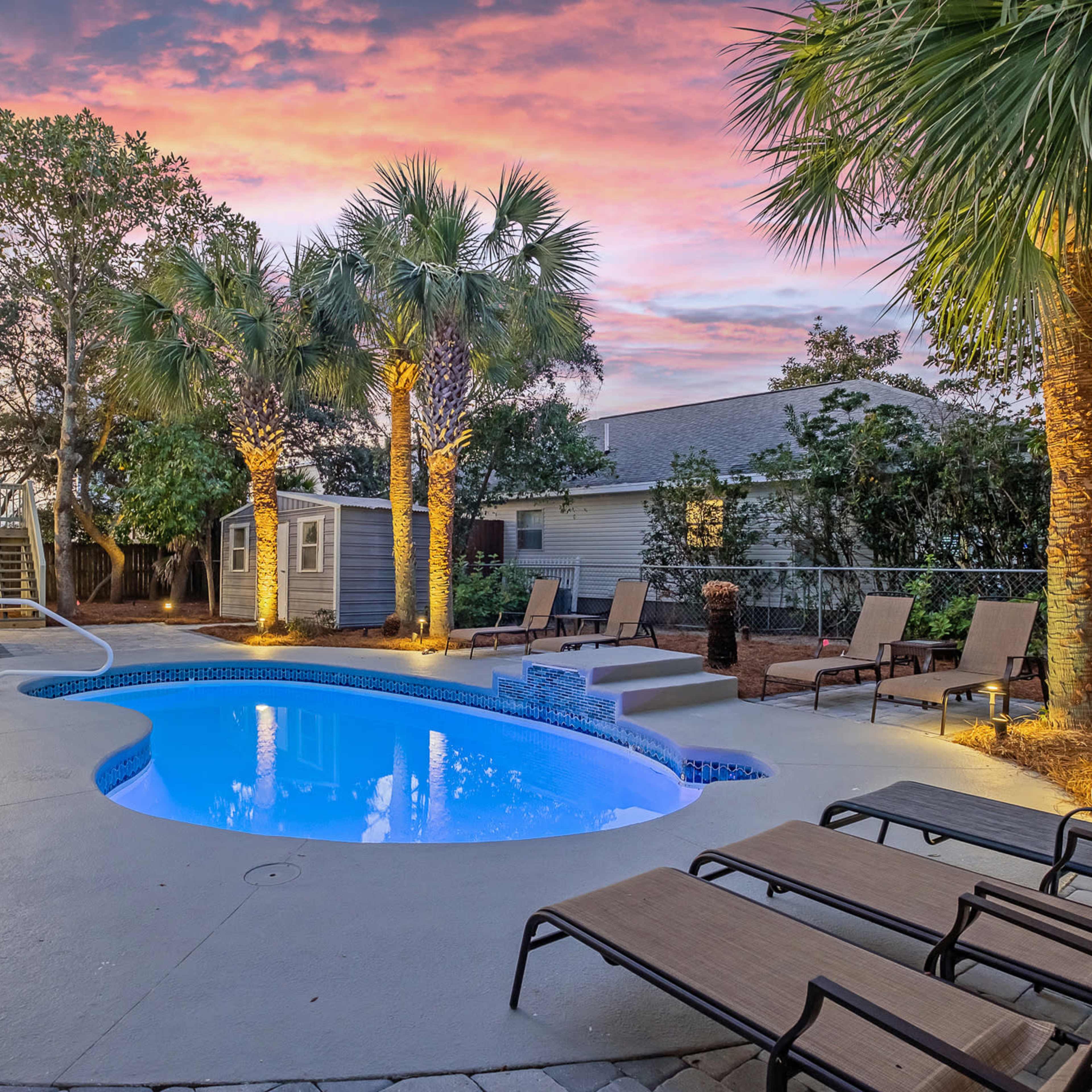 A backyard with a swimming pool surrounded by palm trees and lounge chairs, set against a colorful sunset sky.