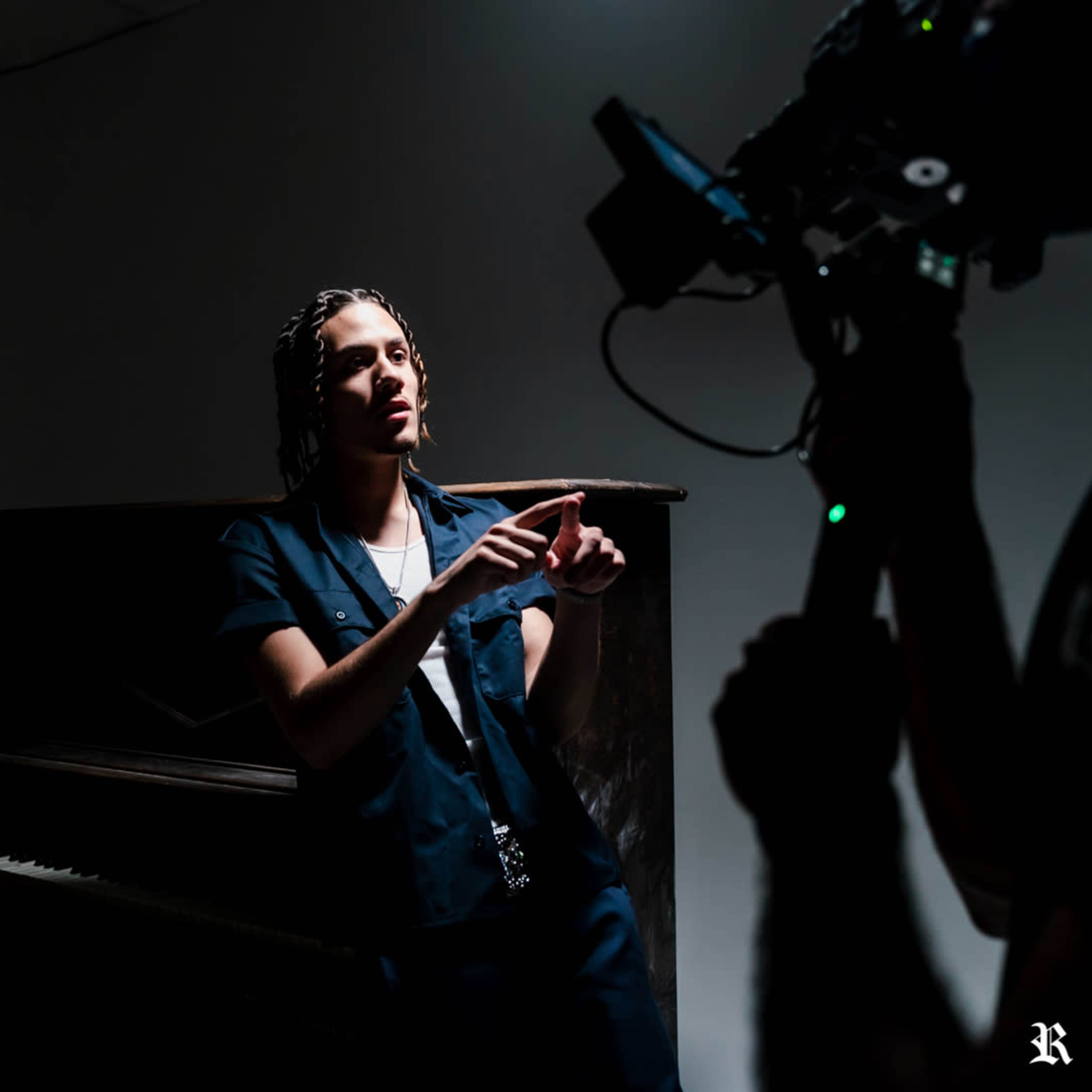 A person in a blue outfit gestures while standing near a piano in a dimly lit studio, with a camera focused on them.
