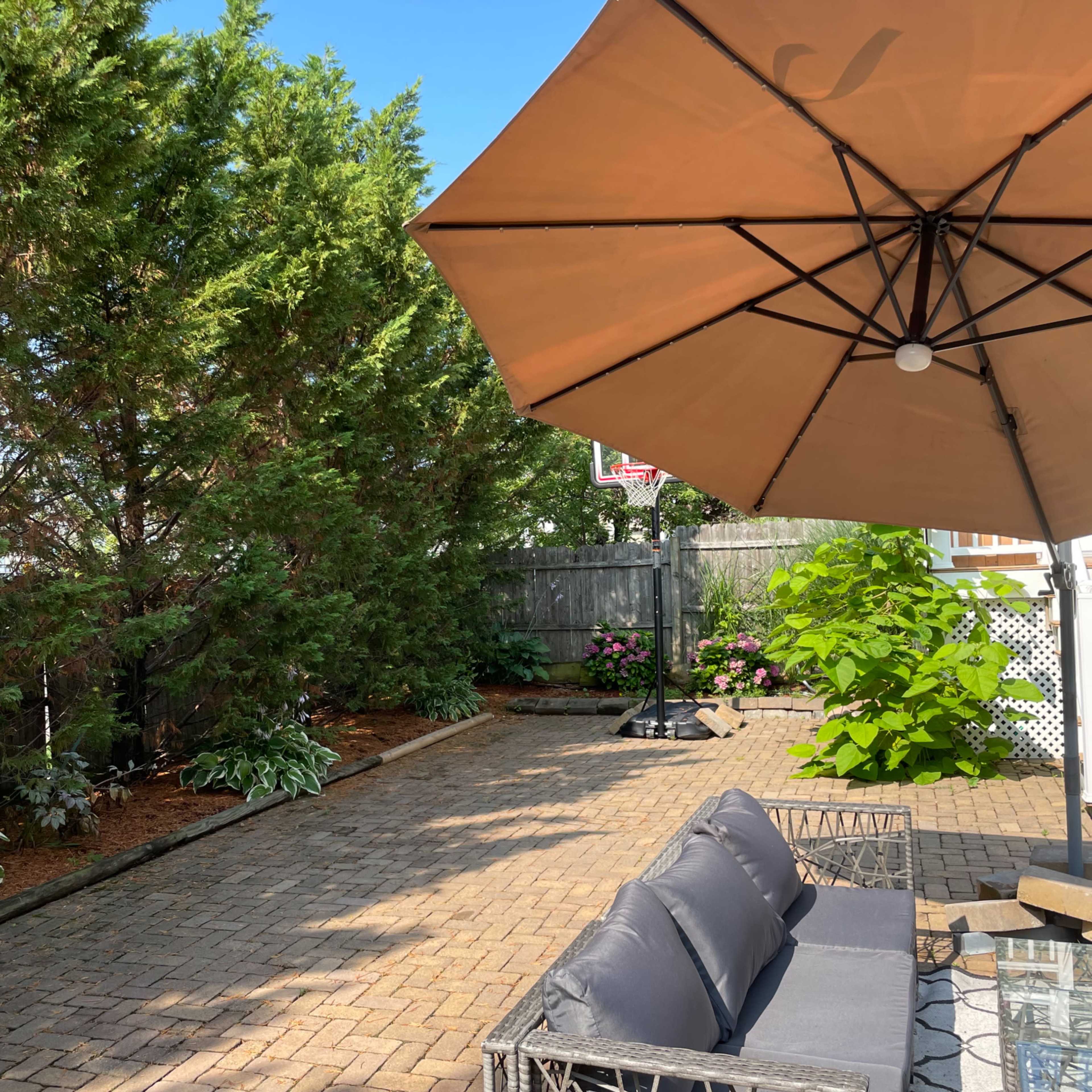 A patio area features a shaded seating arrangement under a large umbrella, surrounded by greenery and flowerbeds.