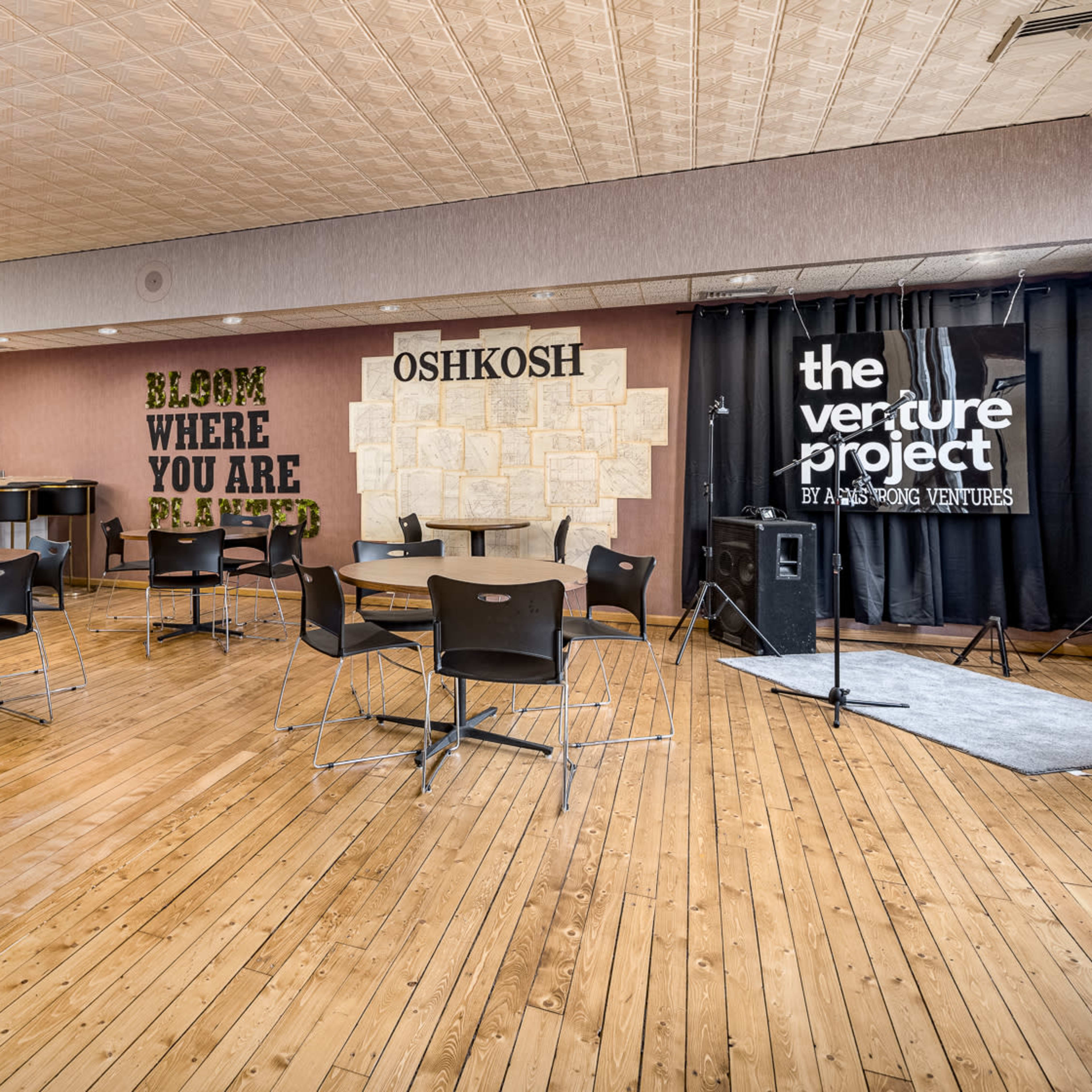 A spacious meeting room features wooden floors, several round tables with black chairs, and a backdrop with decorative text on the walls.