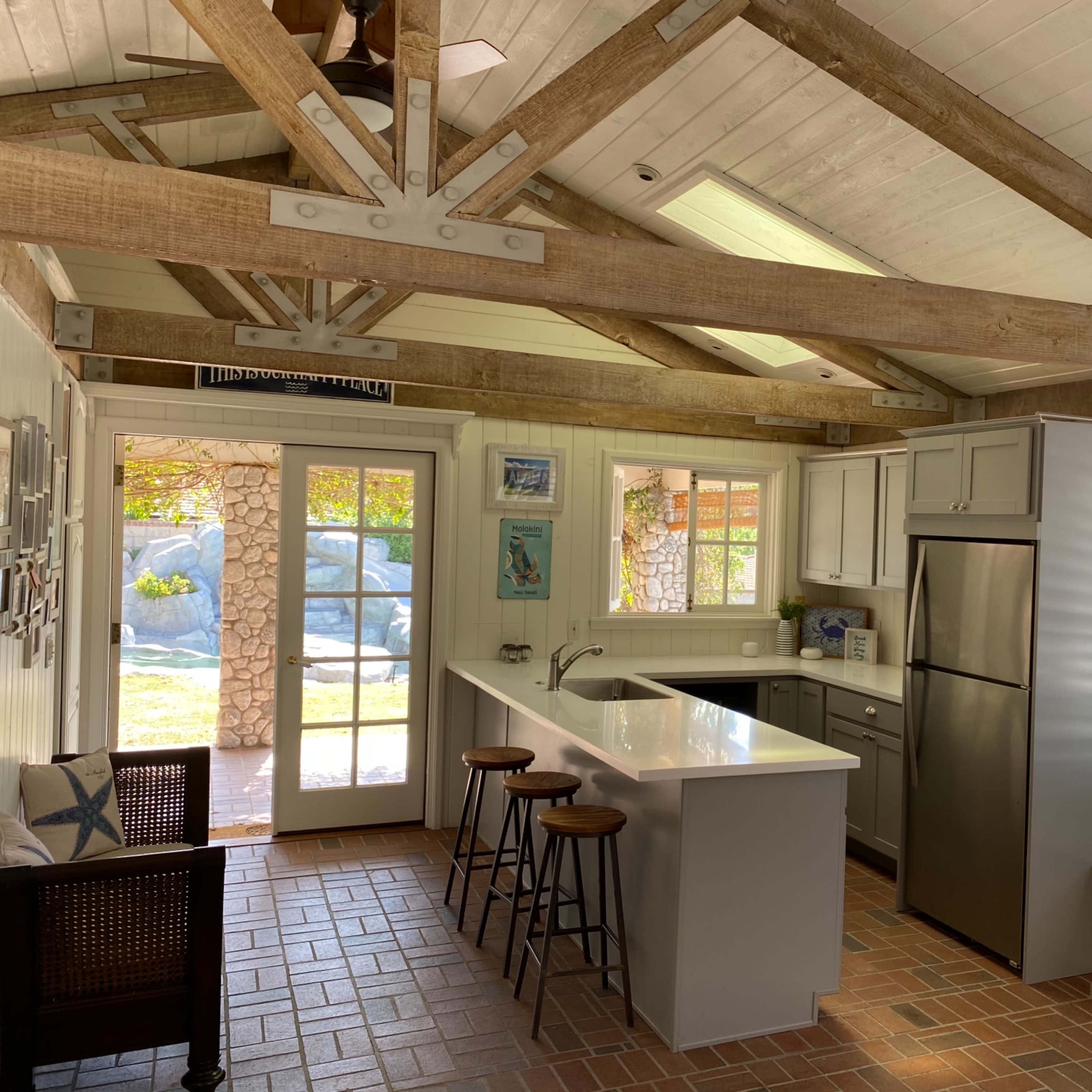A kitchen area with a central island, two bar stools, and wooden beams overhead, featuring light-colored cabinets and a stainless steel refrigerator.