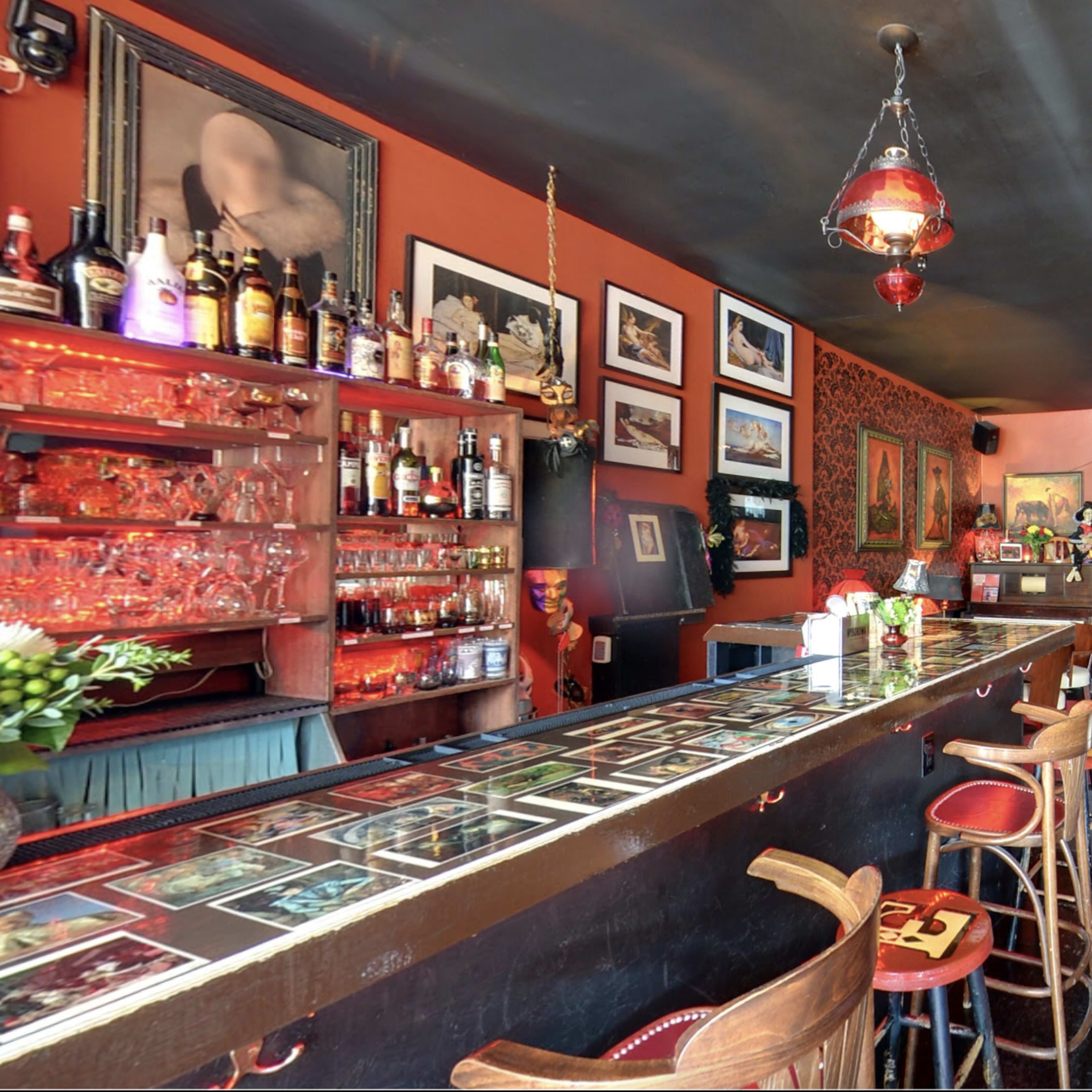 A dimly lit bar with a long counter lined with bottles, surrounded by framed artworks and a few seating areas.