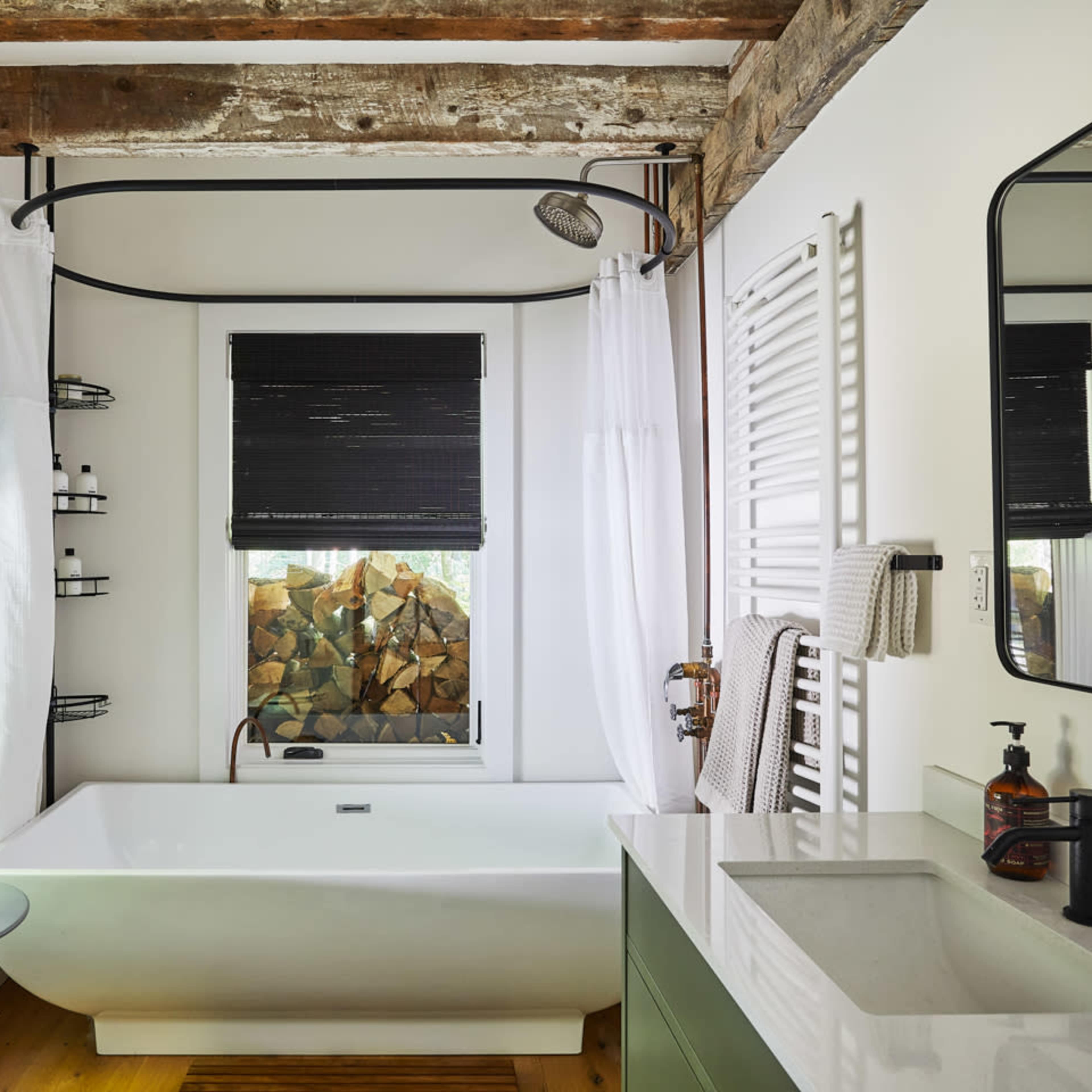The bathroom features a freestanding tub, a modern sink with a mirror, and a window displaying stacked firewood, complemented by wooden beams and contemporary window coverings.