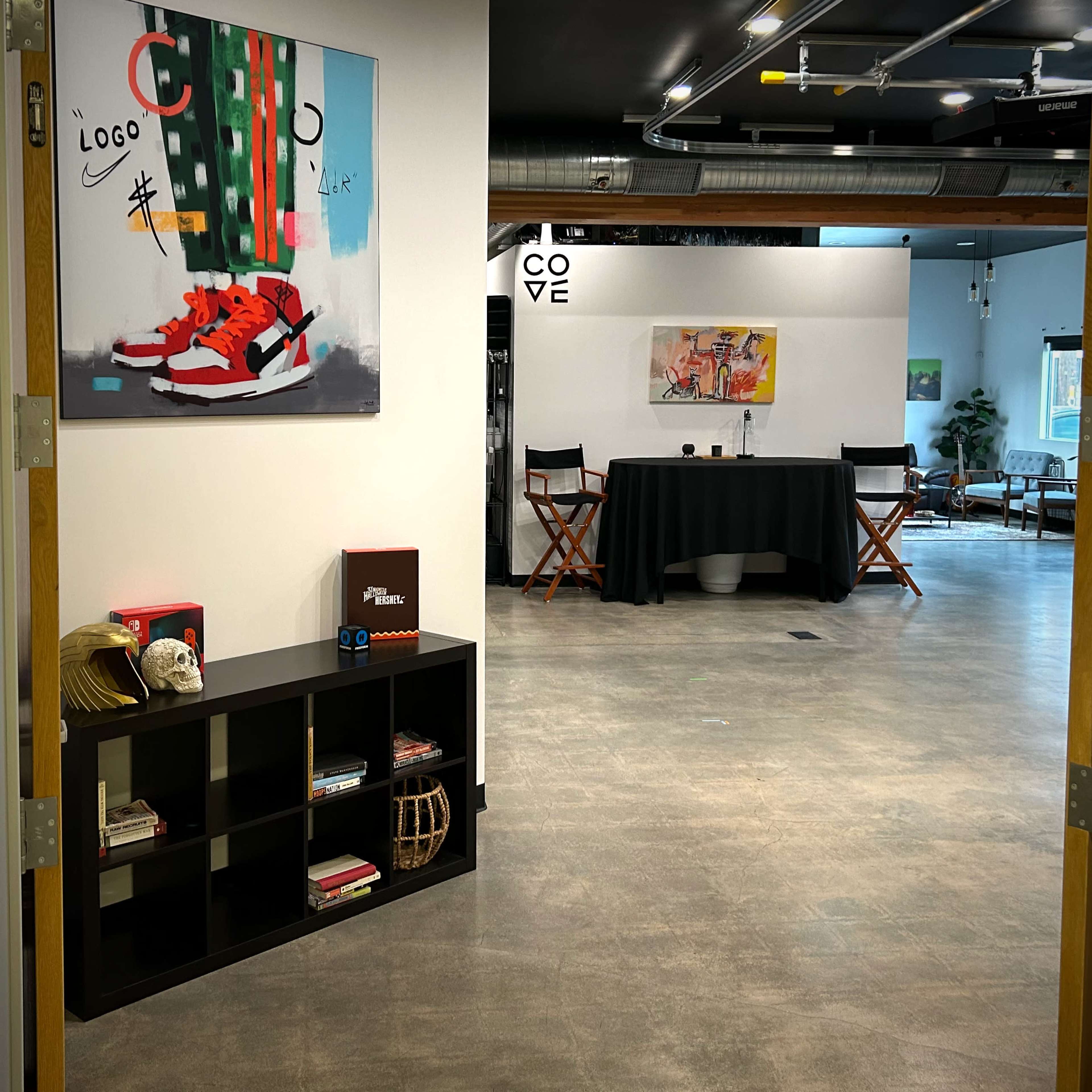 A hallway leads to a spacious room featuring a black table, chairs, and artwork on the walls.