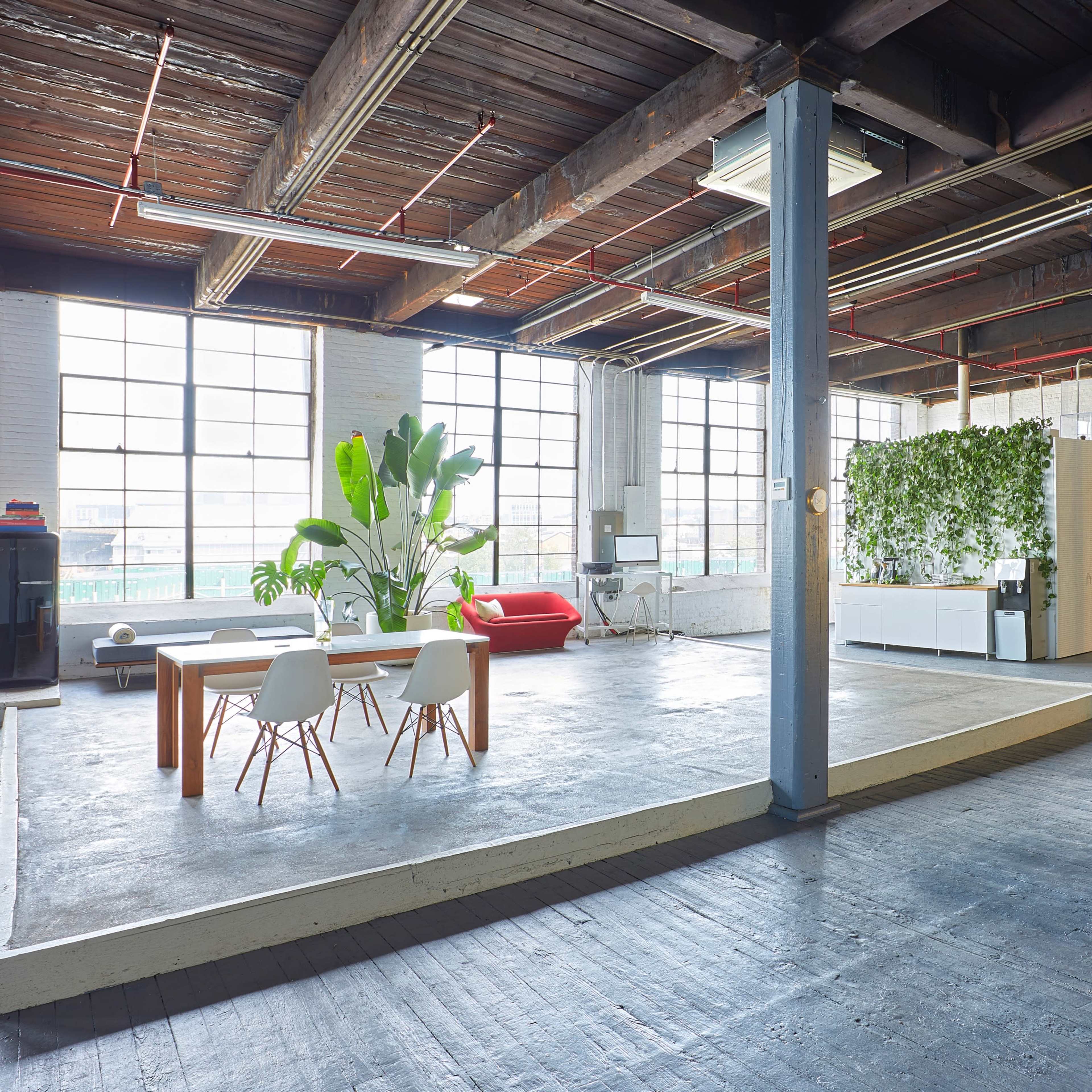 A spacious industrial-style open office with large windows, a dining table with chairs, and green plants integrated into the decor.