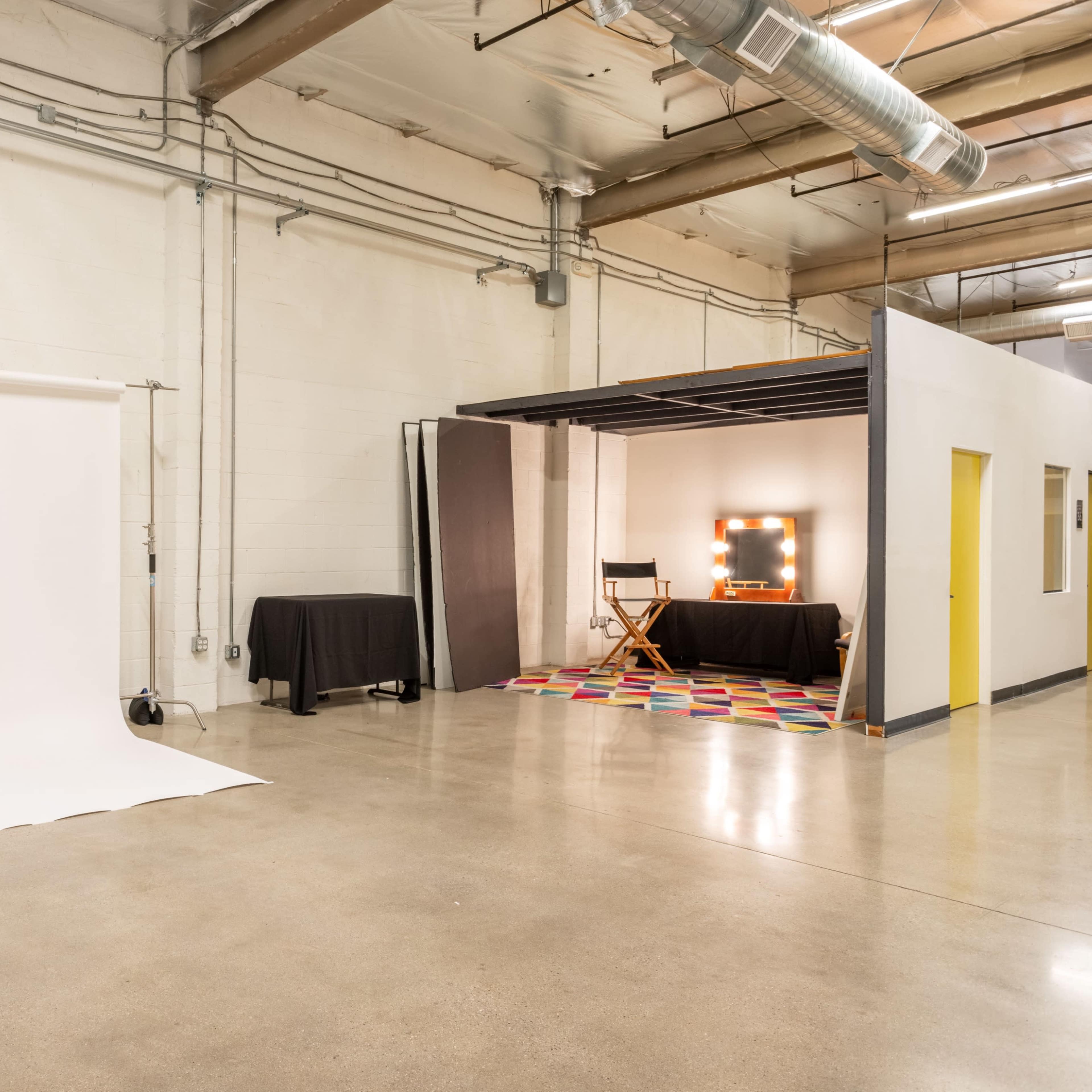 A minimalist studio space with a white backdrop on one side and a styled corner featuring a mirror with lights, a director's chair, and a colorful rug on the other.