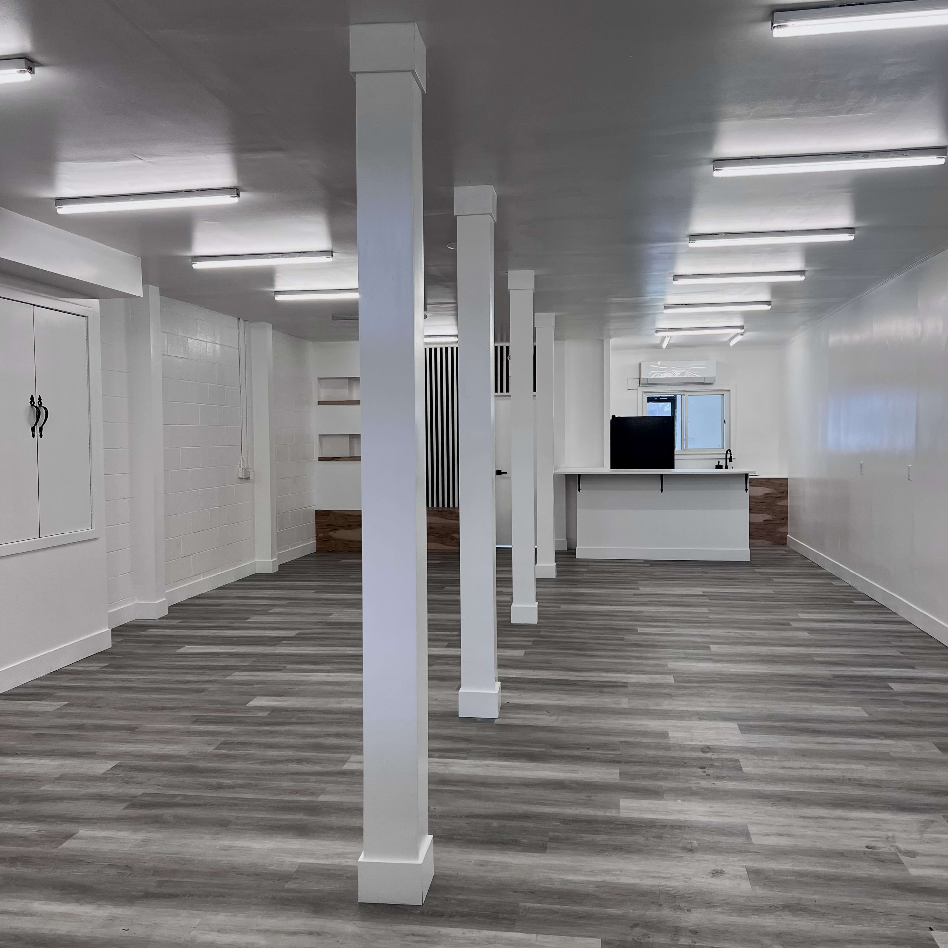 An empty room with gray wooden flooring, white walls, and several white supporting columns, featuring a kitchen area in the back.
