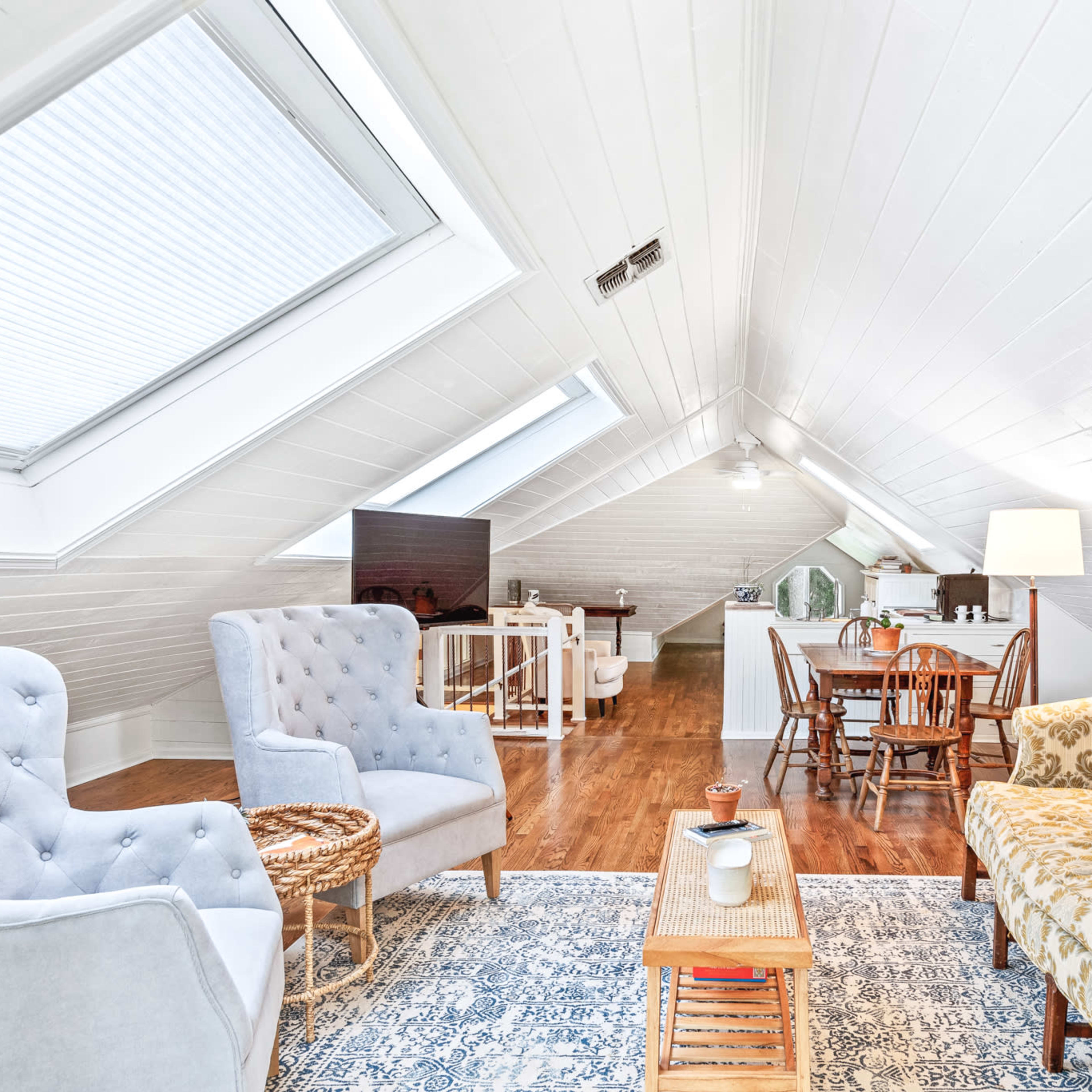 An airy attic space features a cozy seating area with two armchairs and a patterned sofa, alongside a dining table and kitchenette.
