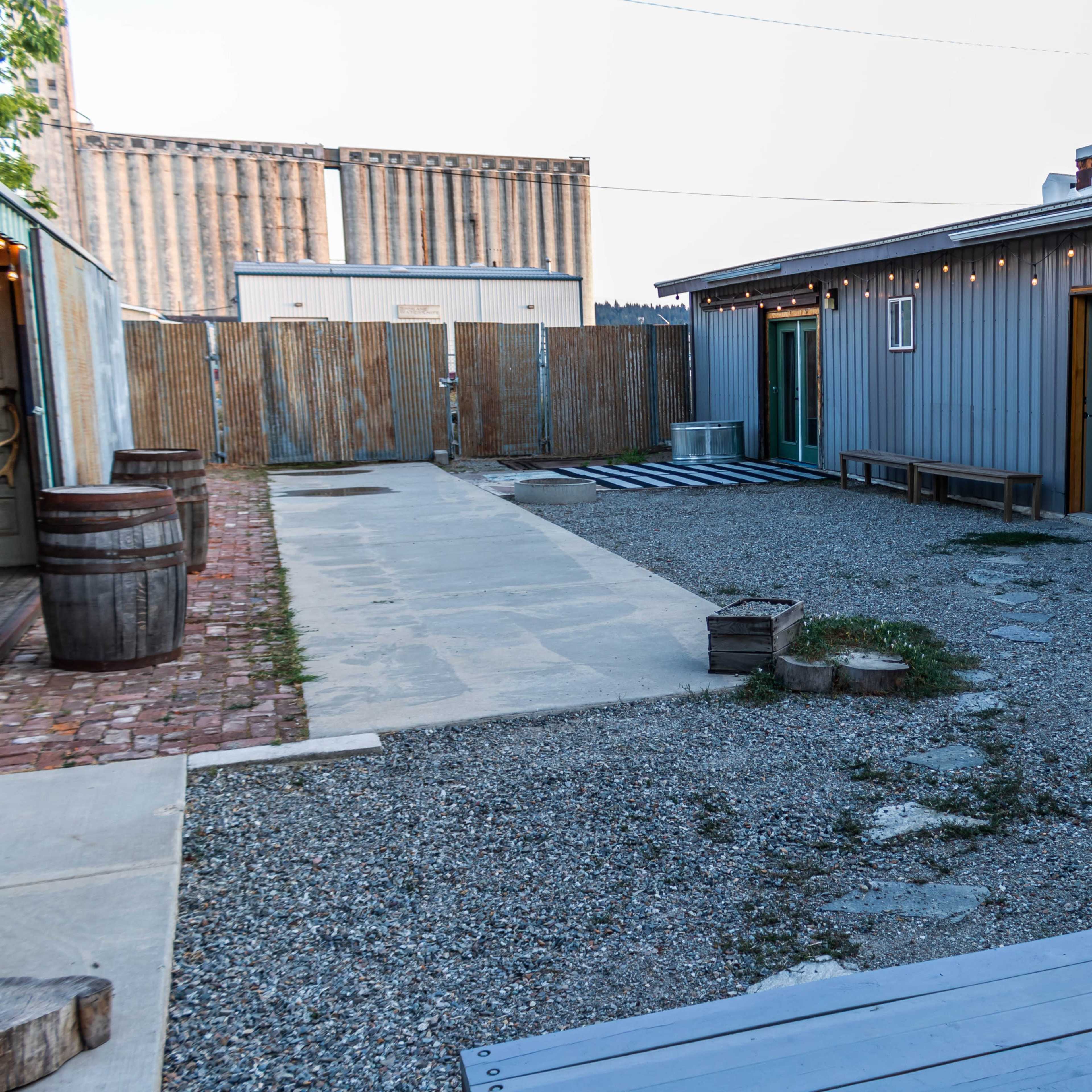 A gravel courtyard surrounded by wooden buildings, with a few benches and barrels positioned to the sides.