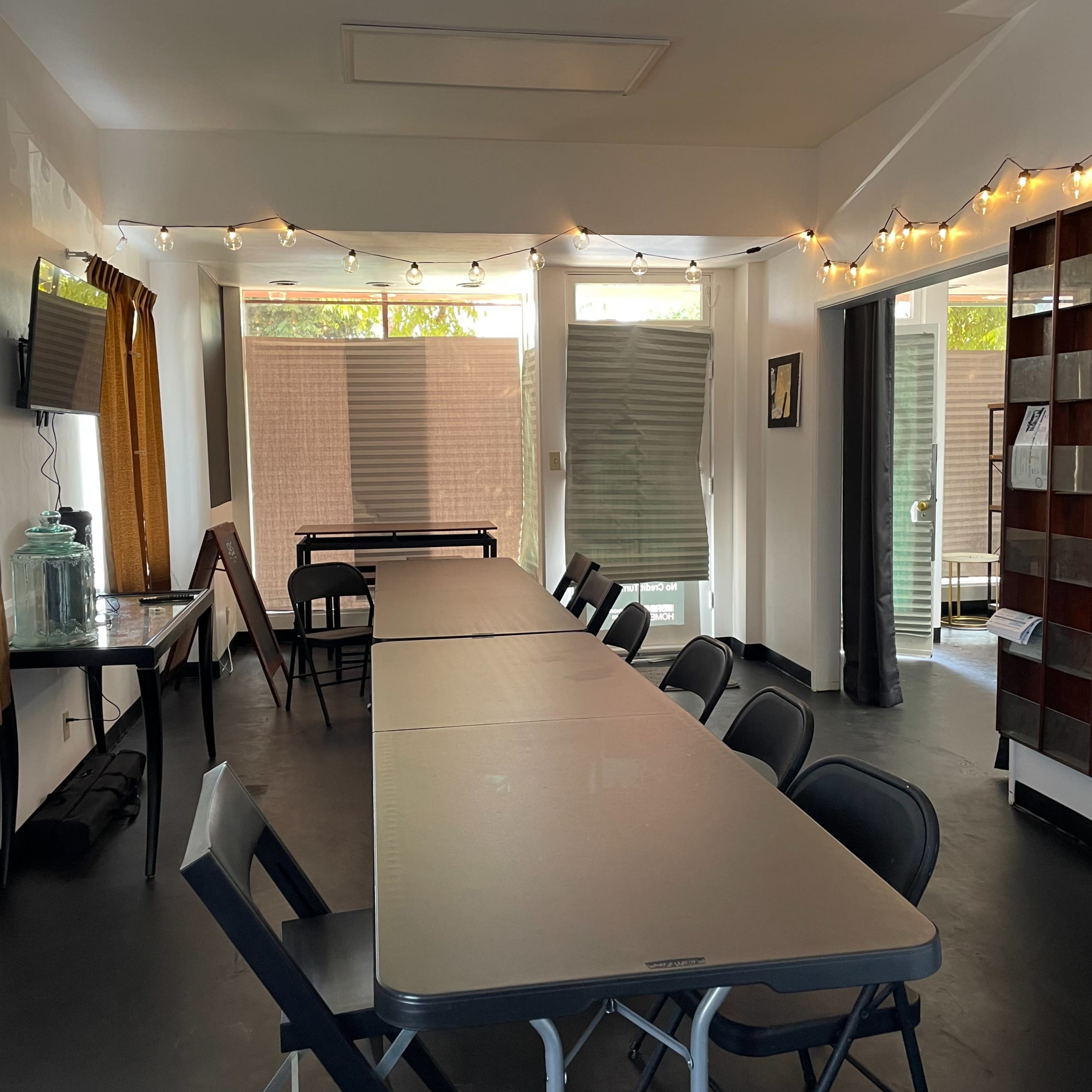 A room with a long table set up with folding chairs, string lights along the walls, and large windows partially covered by blinds.
