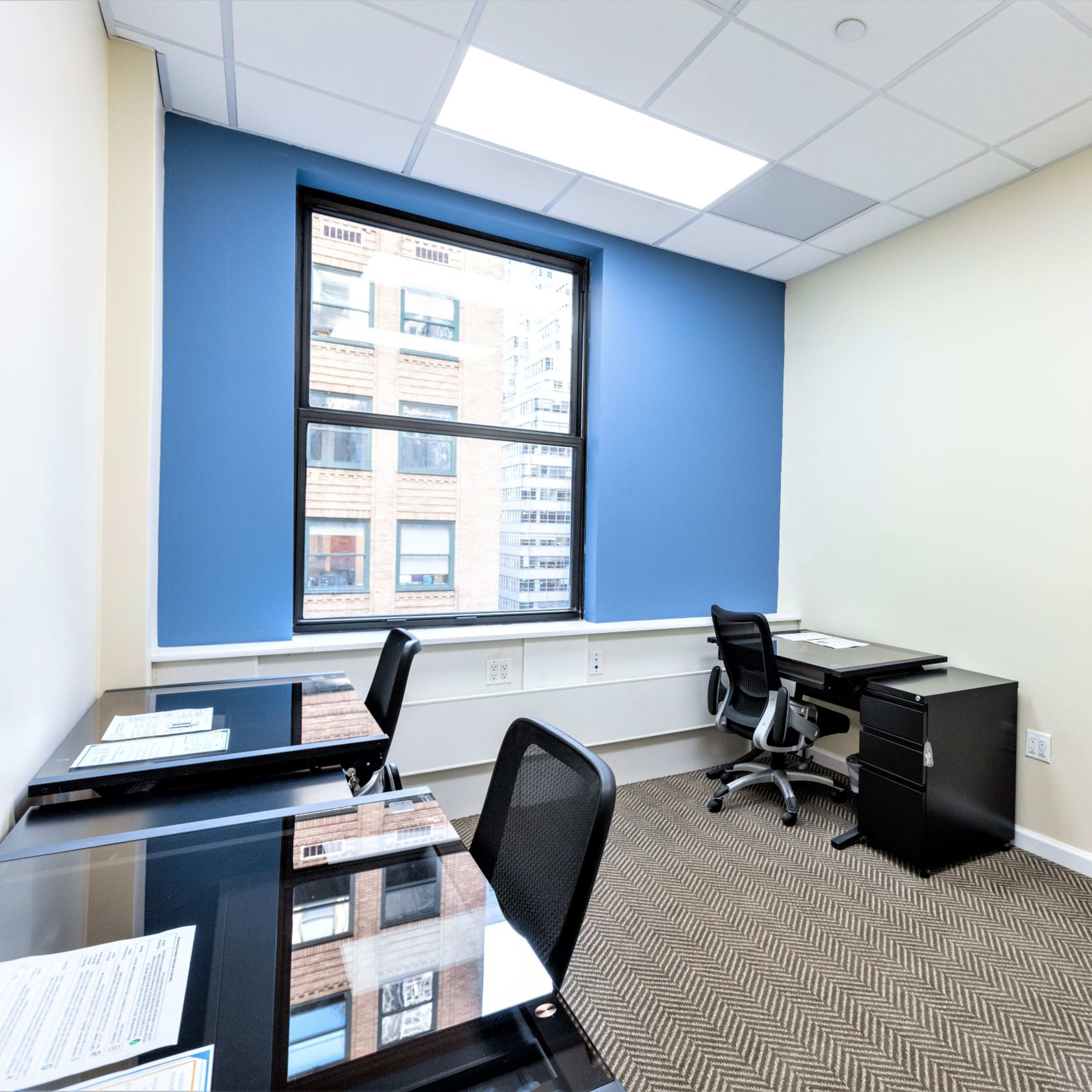 A small office space with two desk setups, a black office chair, and a large window letting in natural light.