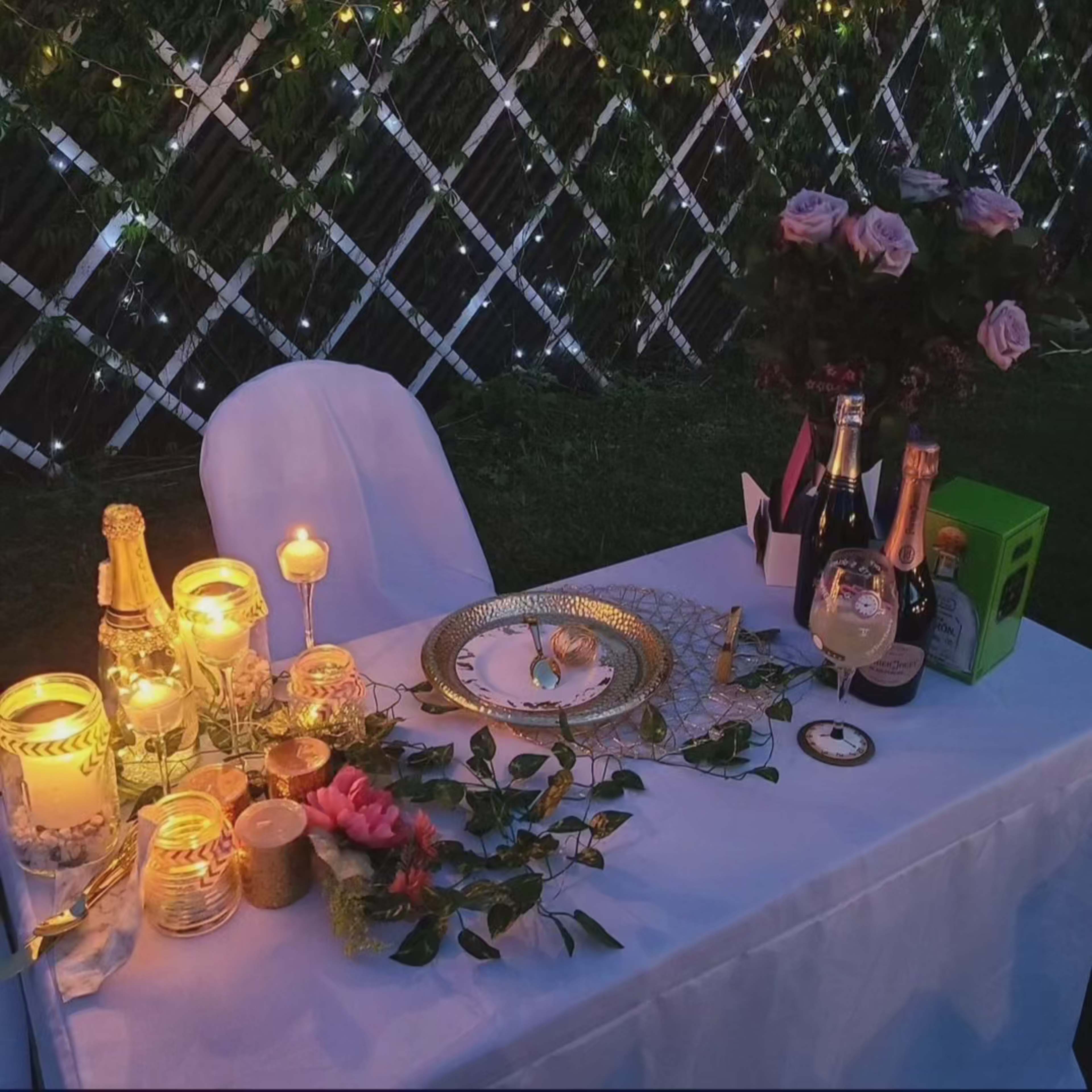A decorated table for two is set with candles, champagne bottles, and flowers, against a backdrop of illuminated greenery.