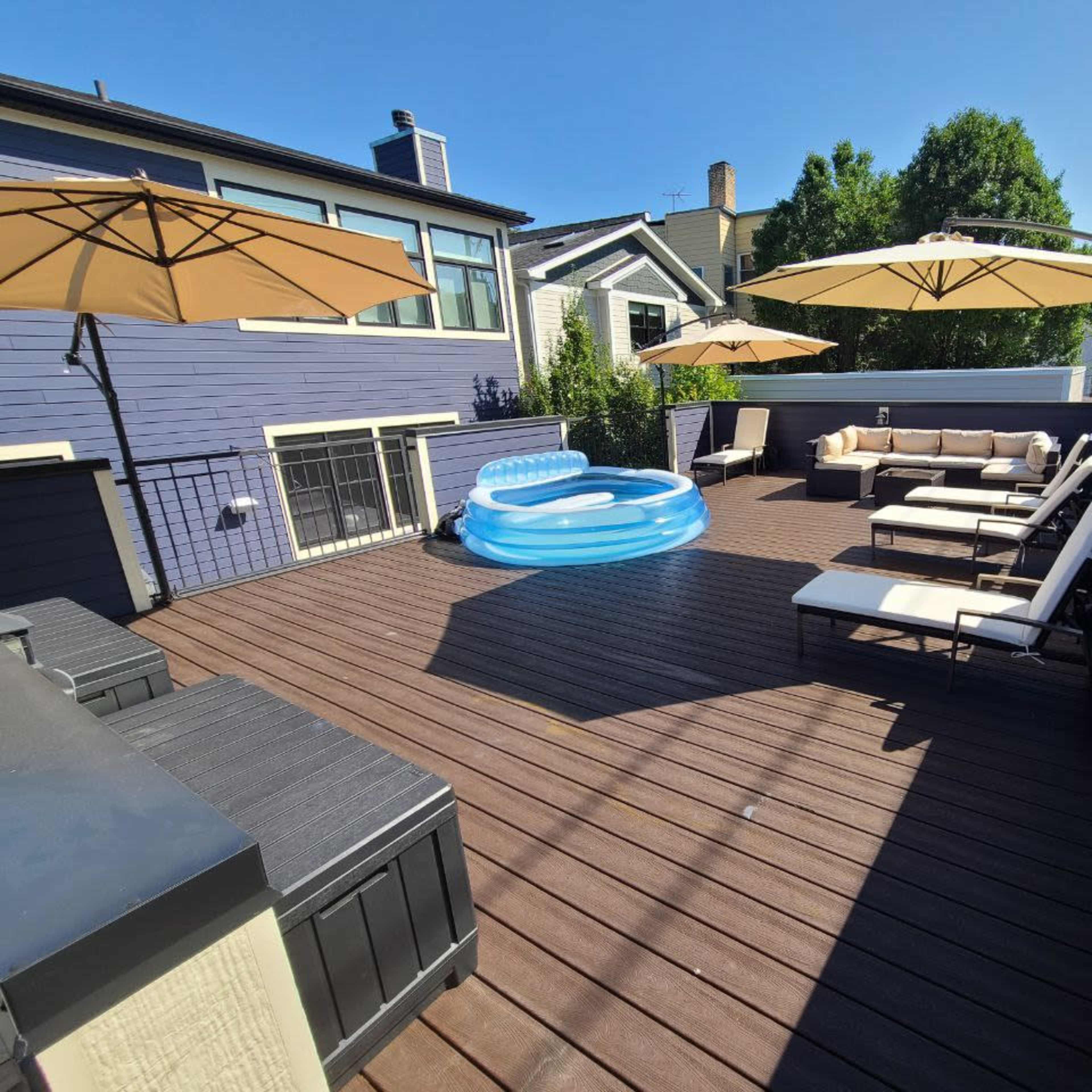 A spacious wooden deck equipped with lounge chairs, a beige sectional sofa, a circular inflatable pool, and several umbrellas for shade.