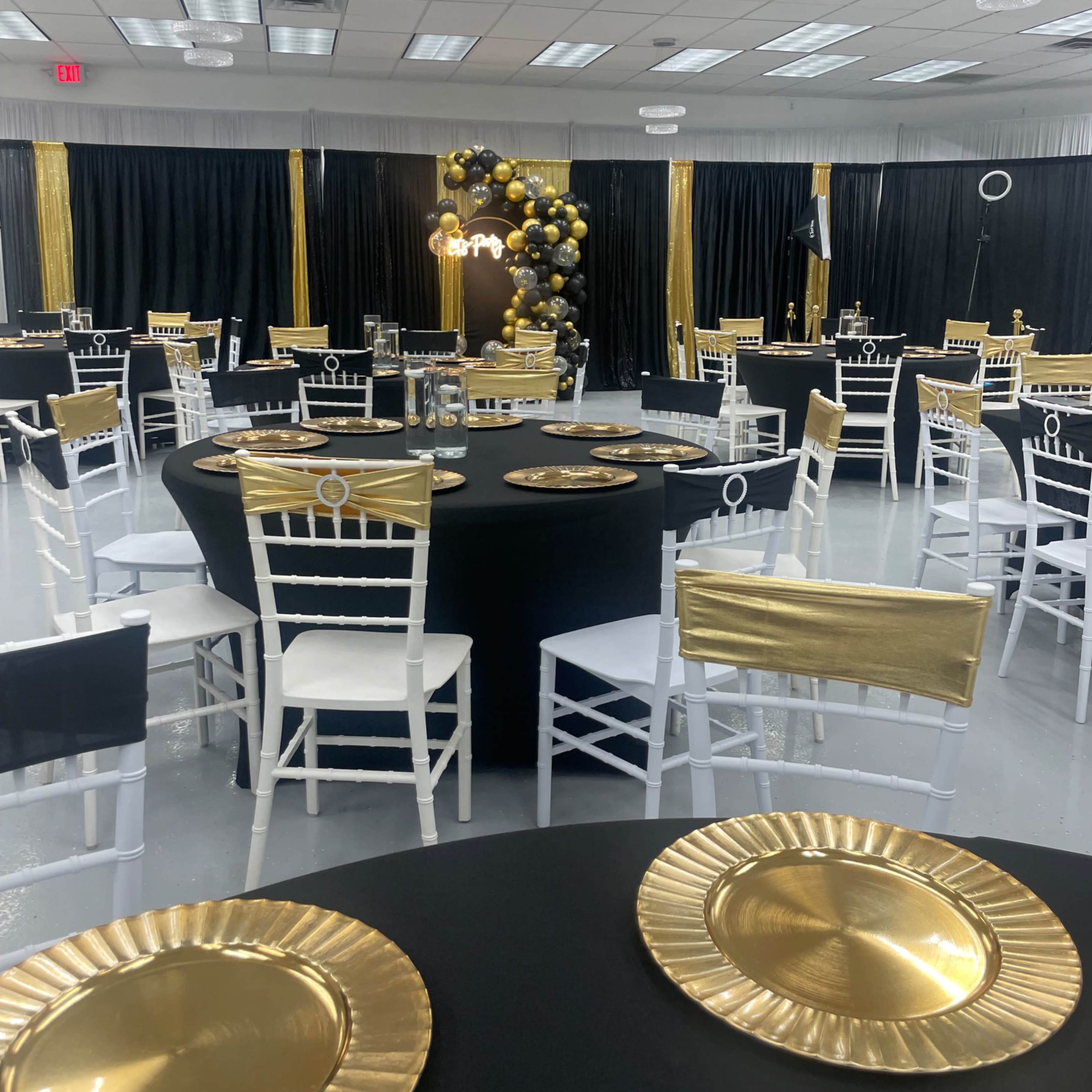 A banquet hall is set up with black and gold decor, featuring round black tables, white chairs, and decorative gold plates.