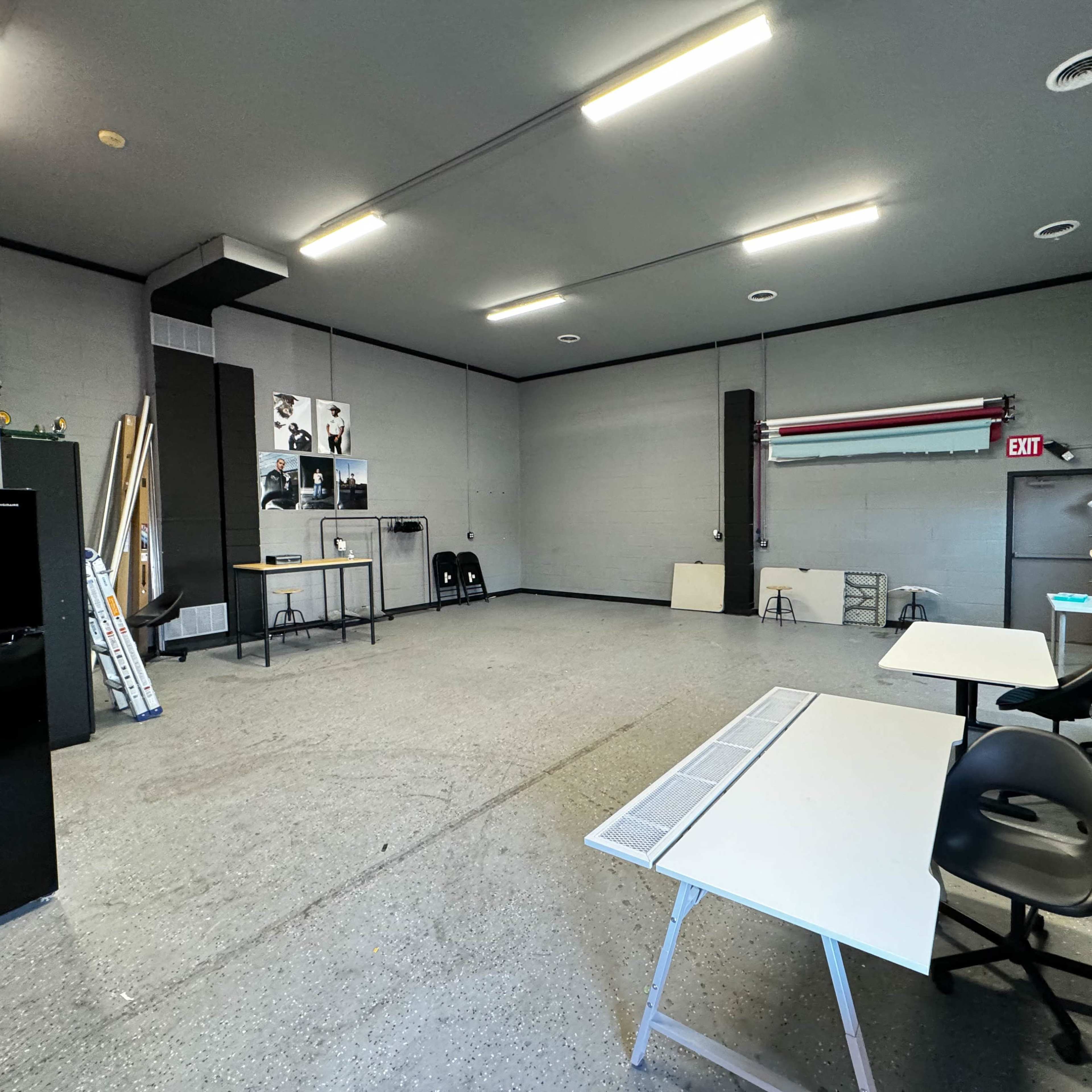A spacious, empty room with grey walls, two tables, a black refrigerator, and light fixtures on the ceiling.