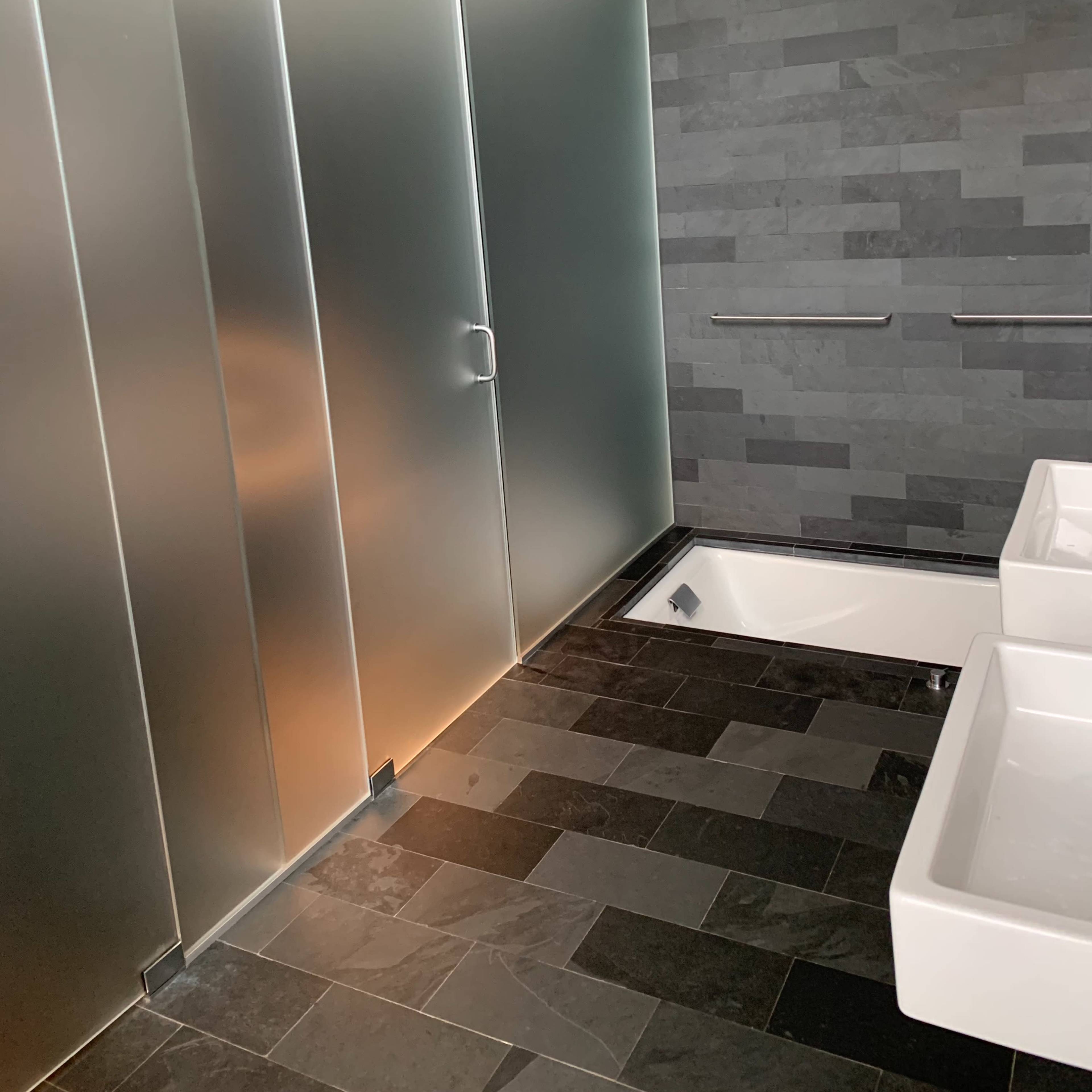 A modern bathroom features frosted glass doors, a stone wall, and dual sinks on a dark tiled floor.