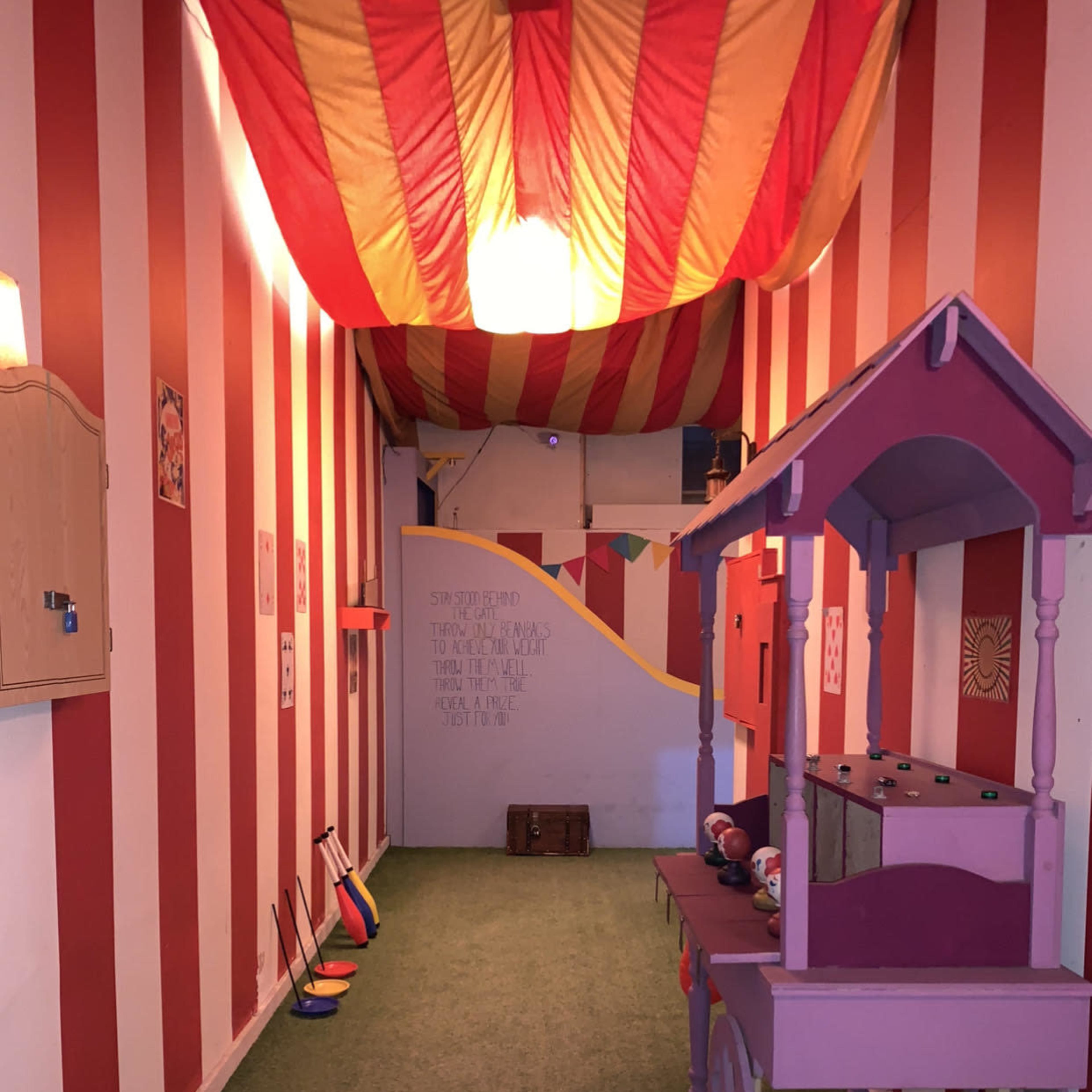 A narrow hallway decorated with red and white striped walls, leading to a colorful play area featuring a pink cart and various toys.