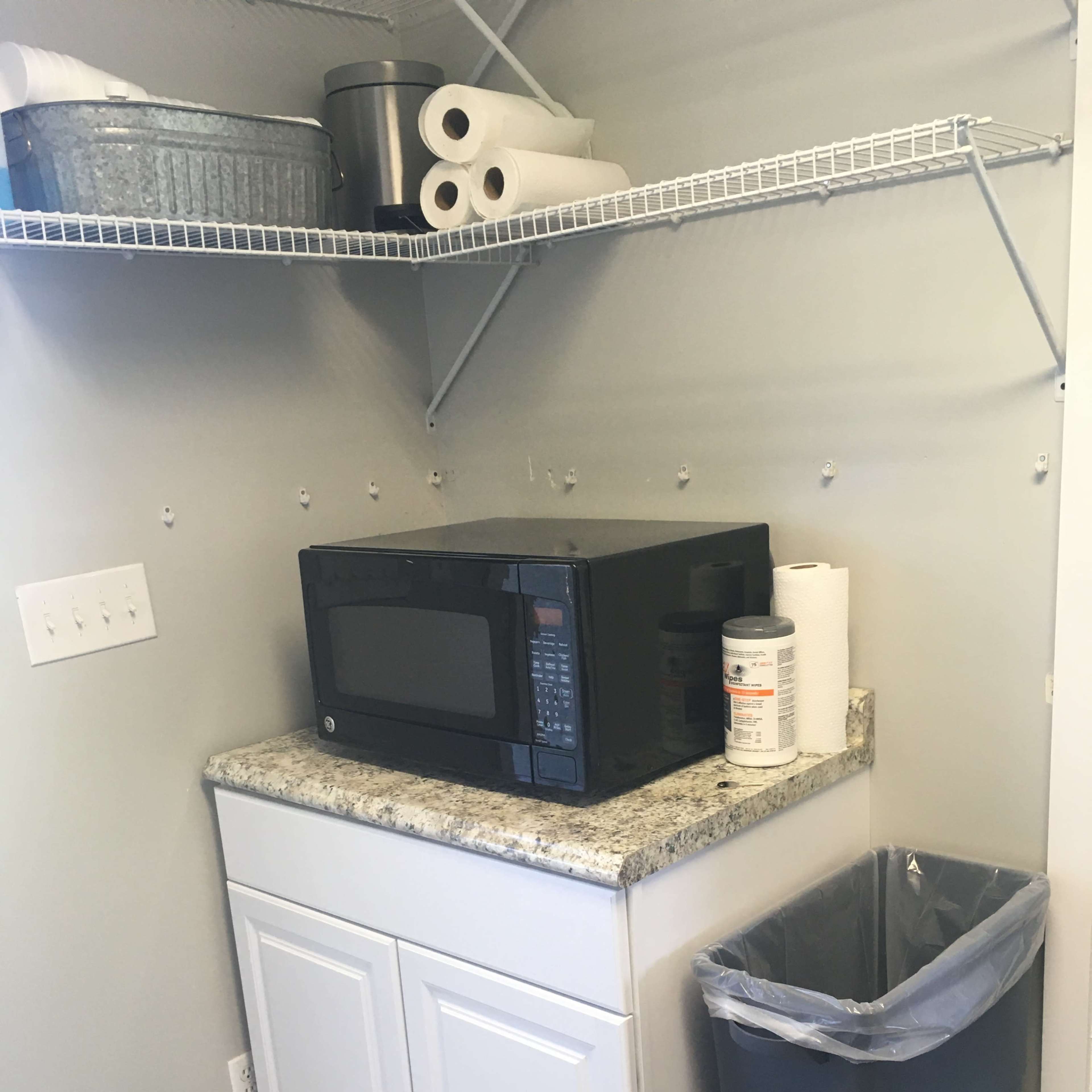 A laundry room with a microwave placed on a granite countertop below a shelf holding various containers and paper towels, alongside a trash bin.