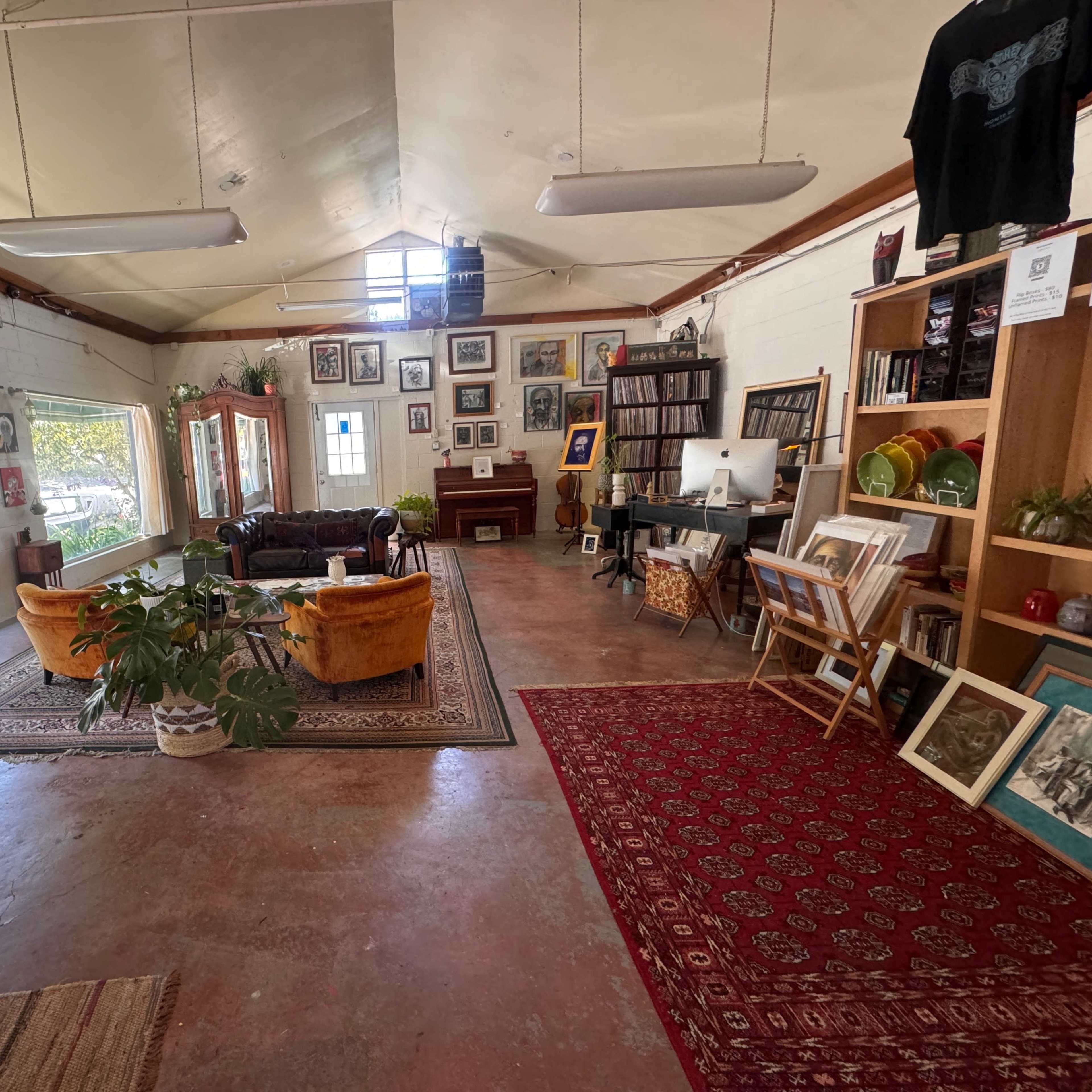 A spacious, well-lit room featuring a blend of seating areas, shelves filled with books and artworks, and various plants, all arranged around a central rug.