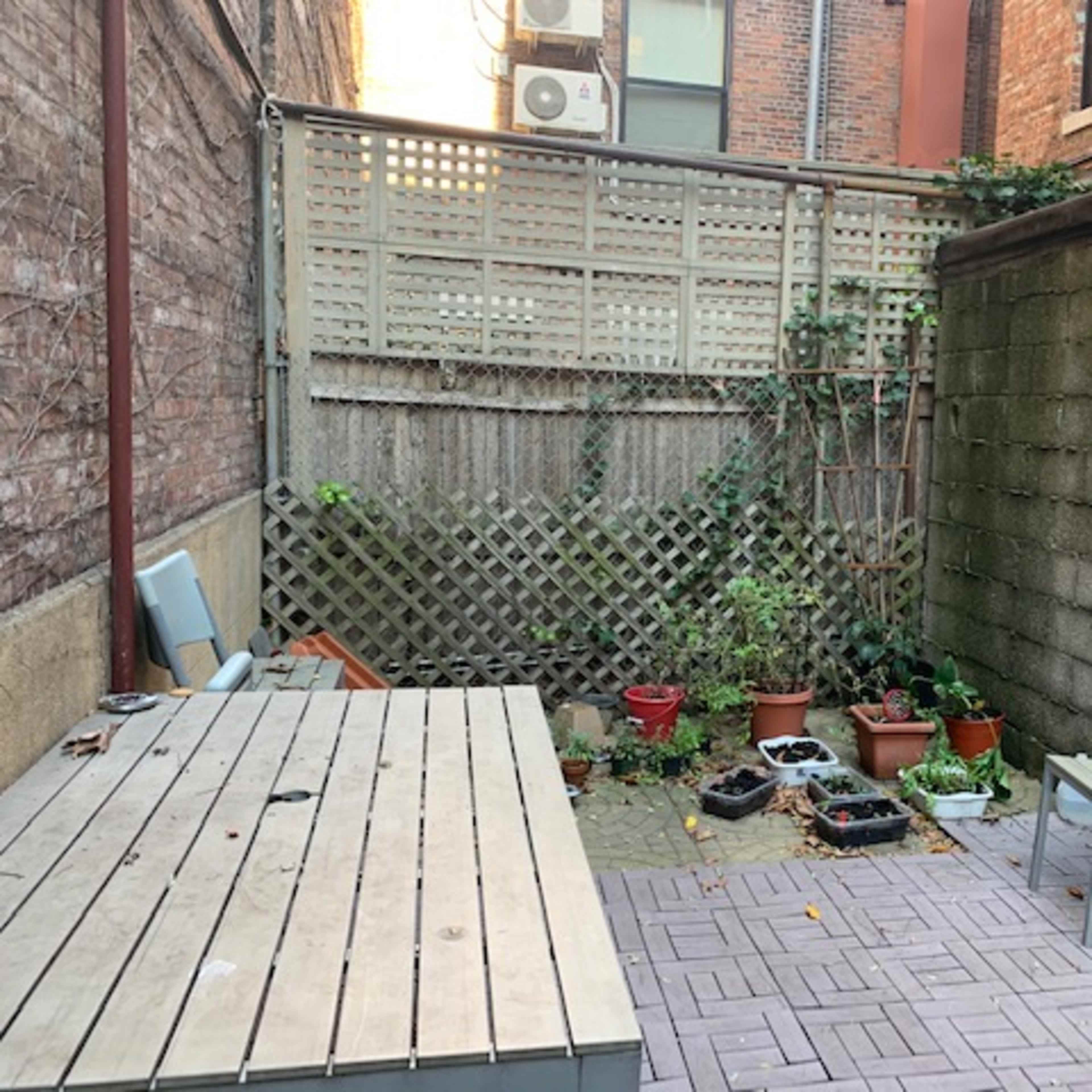 A small brick-paved courtyard with a wooden table, a chair, and several potted plants along the walls.
