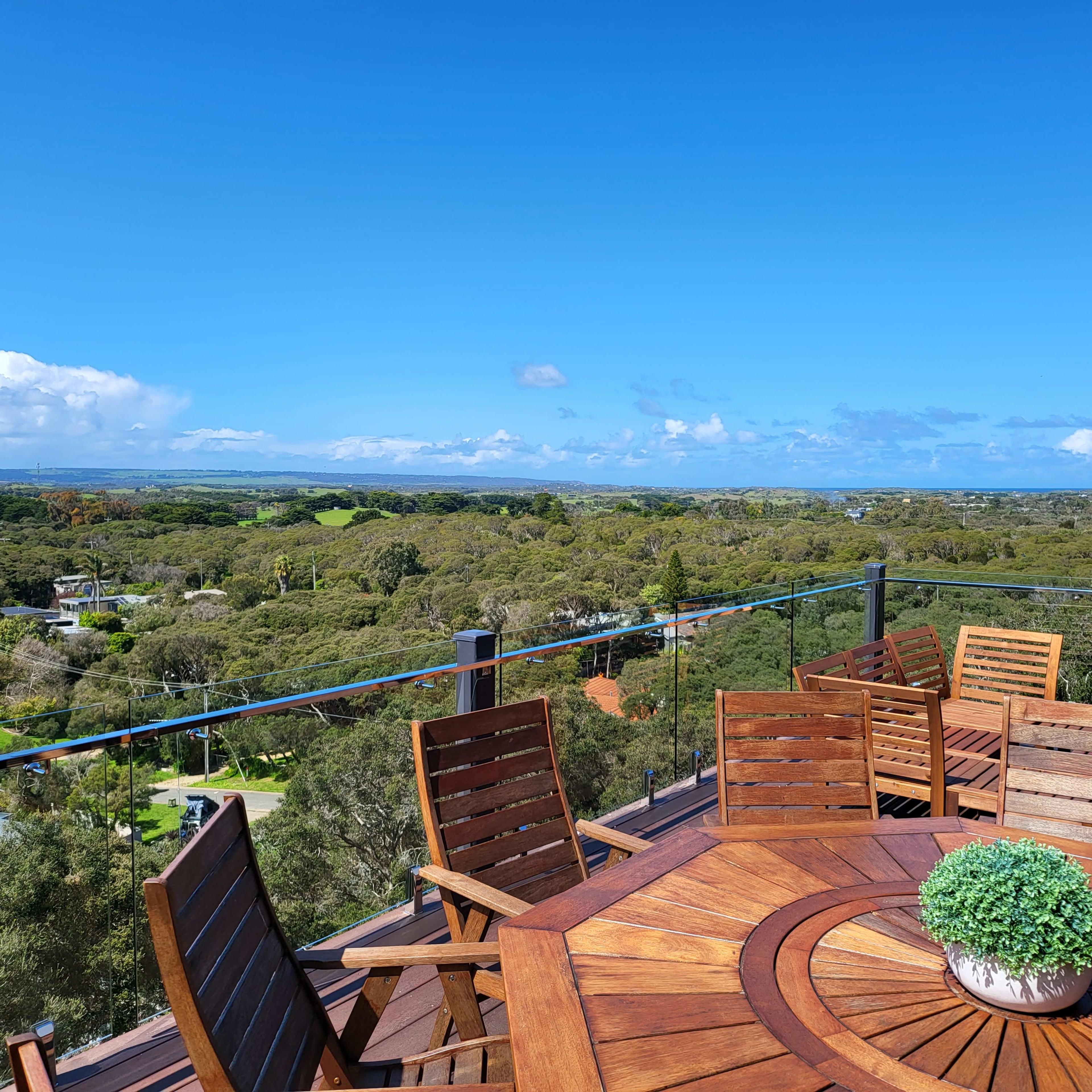 A wooden dining table surrounded by chairs sits on a balcony with a view of green hills and blue skies.