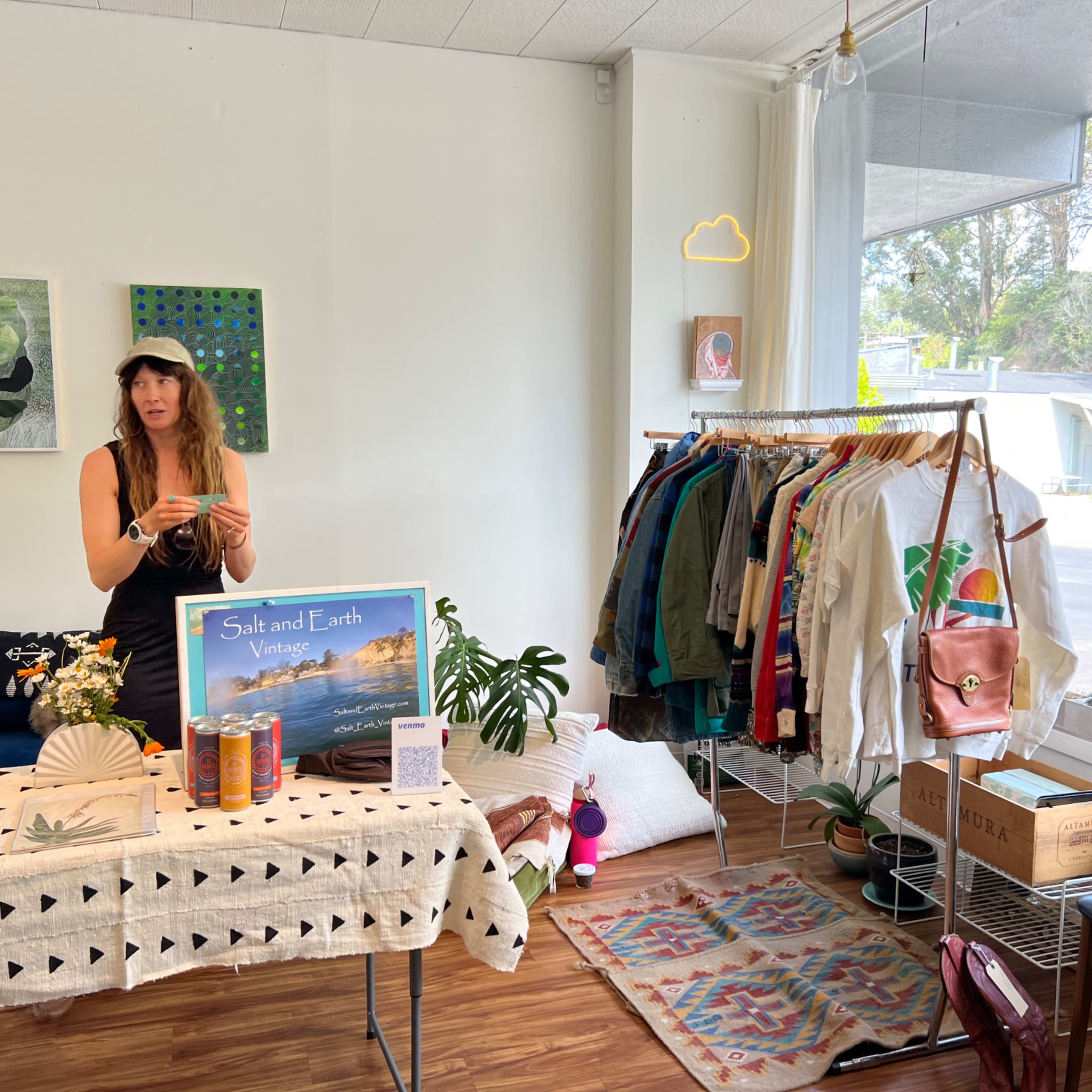 A woman stands behind a display table in a vintage clothing shop, with racks of colorful garments and decorative items surrounding her.