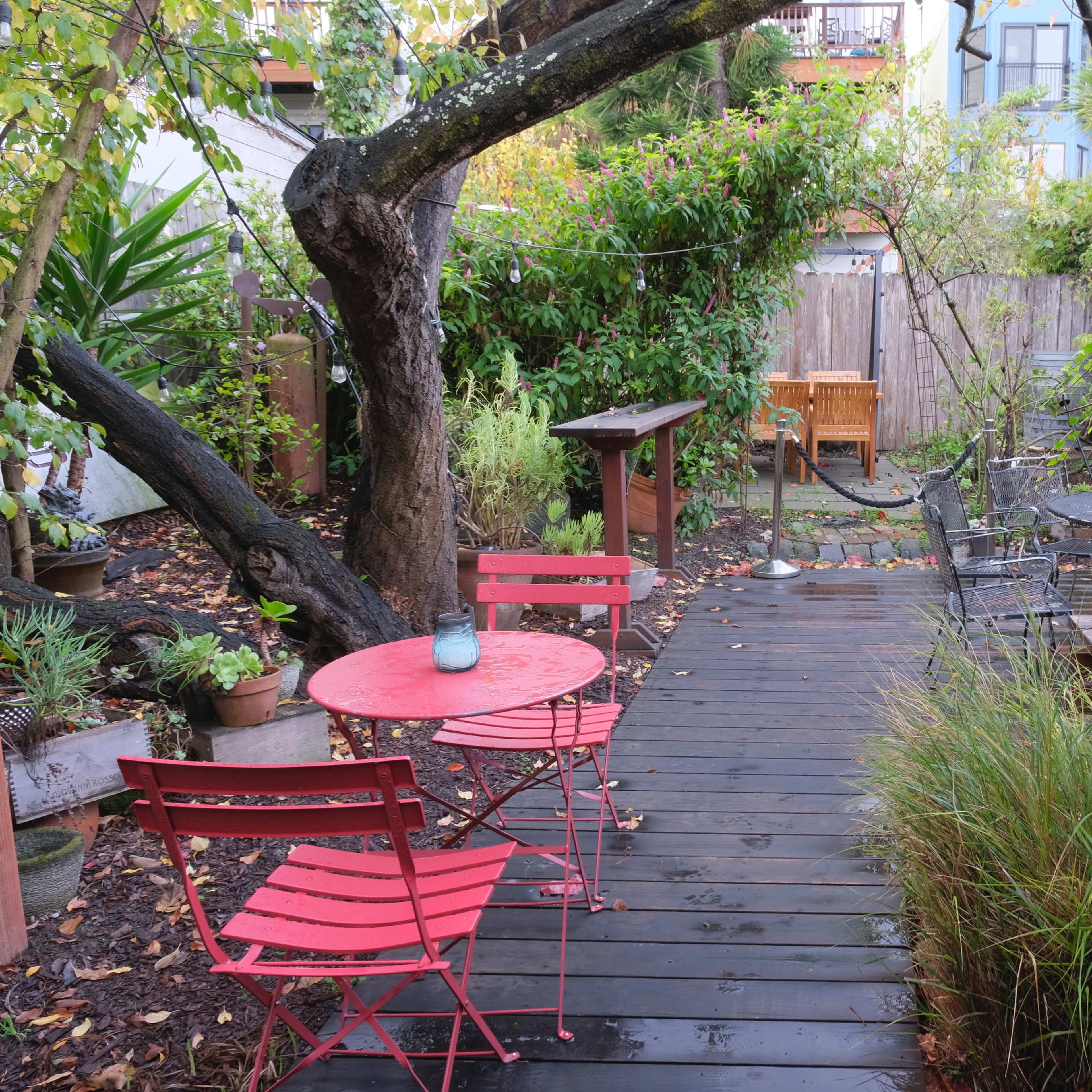 A narrow wooden pathway leads through a garden with potted plants, red chairs and tables, and a wooden seating area in the background.
