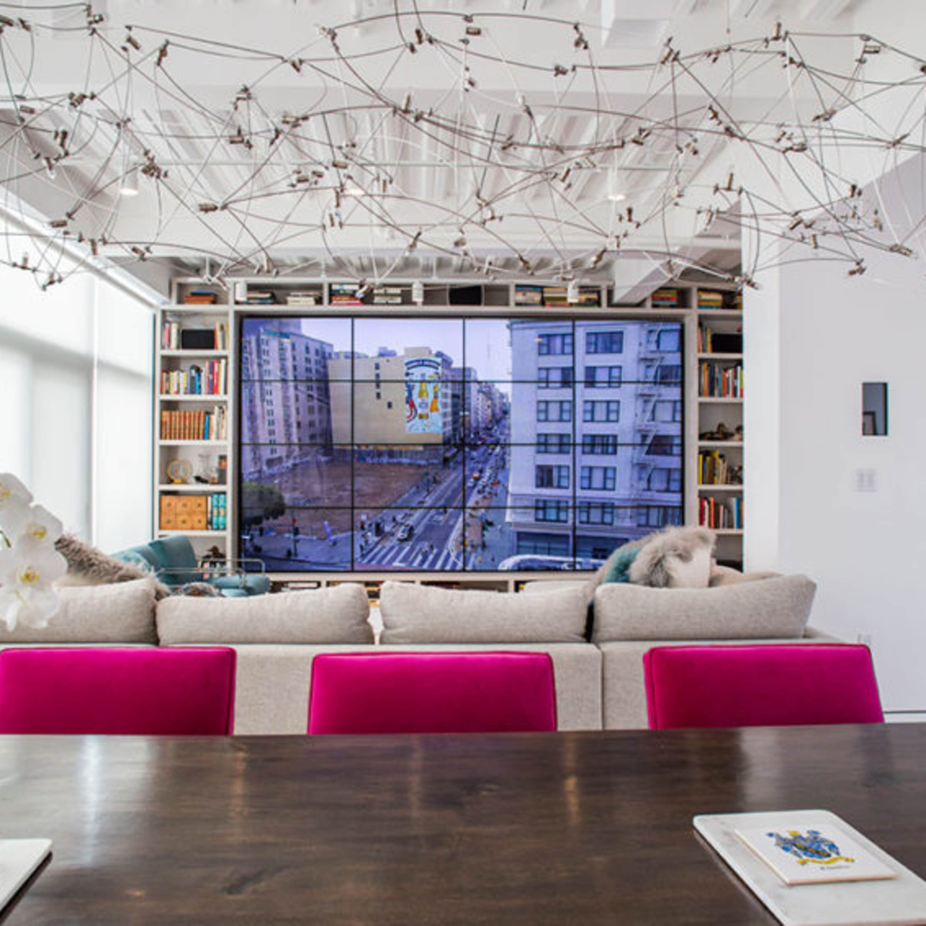 A modern living room features a large television mounted on a white wall, surrounded by bookshelves, with a couch adorned with pink cushions and a dining table in the foreground.