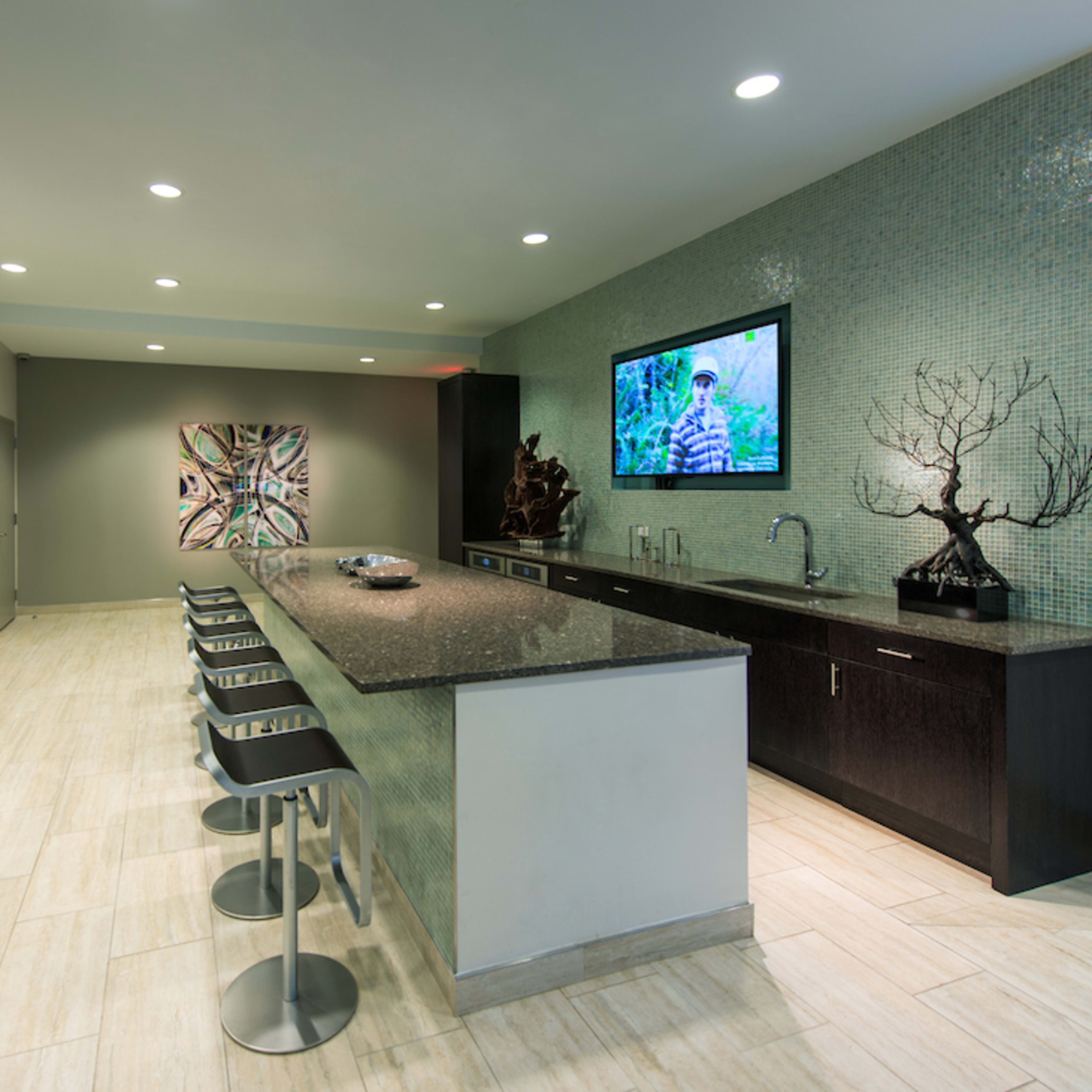 A modern kitchenette with a long granite counter, bar stools, a television on the wall, and artwork displayed on the adjacent wall.
