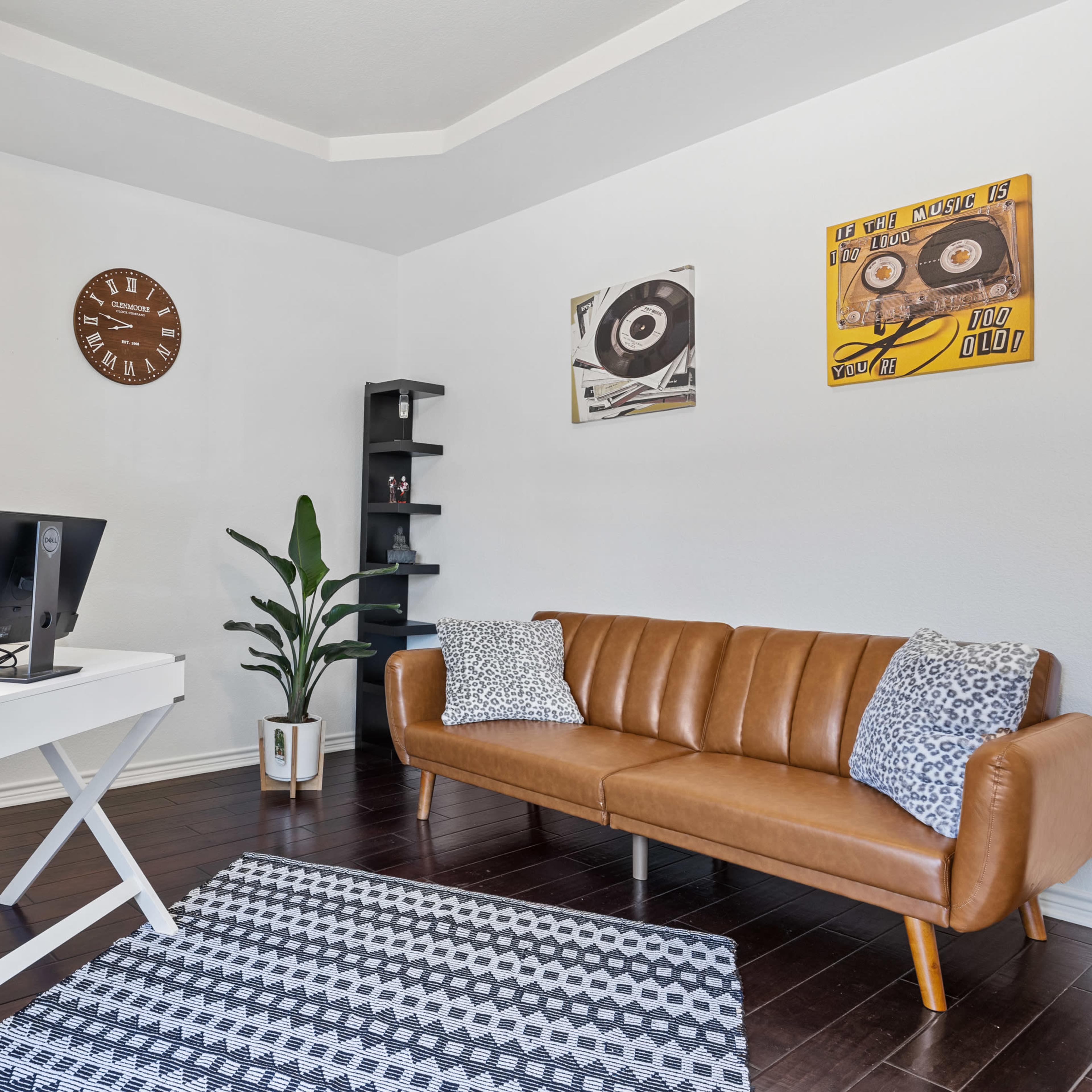 A modern home office with a desk, a computer, an orange chair, a brown leather sofa, and decor including wall art and a clock.
