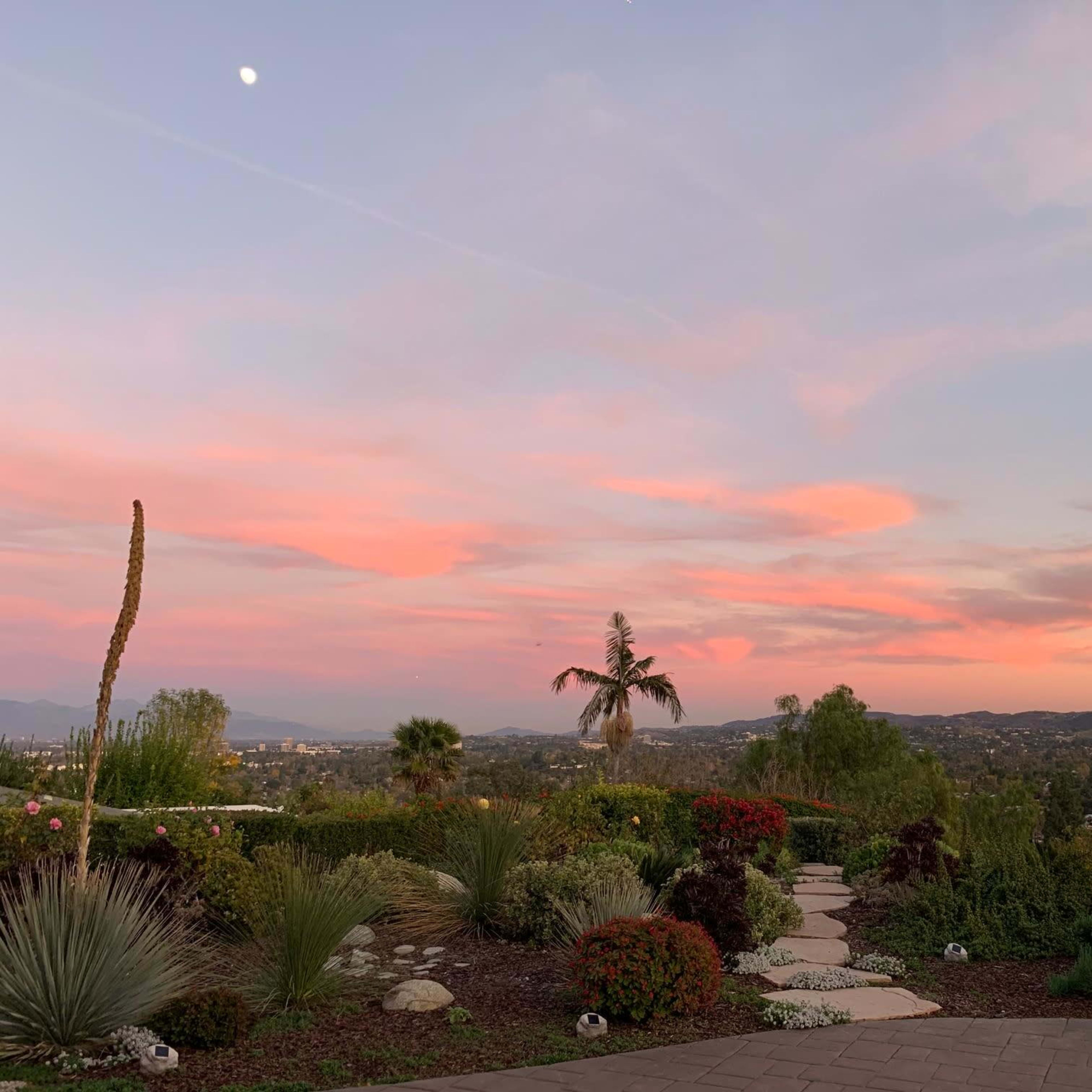 A pathway winds through a landscaped garden under a colorful sunset sky with distant mountains.