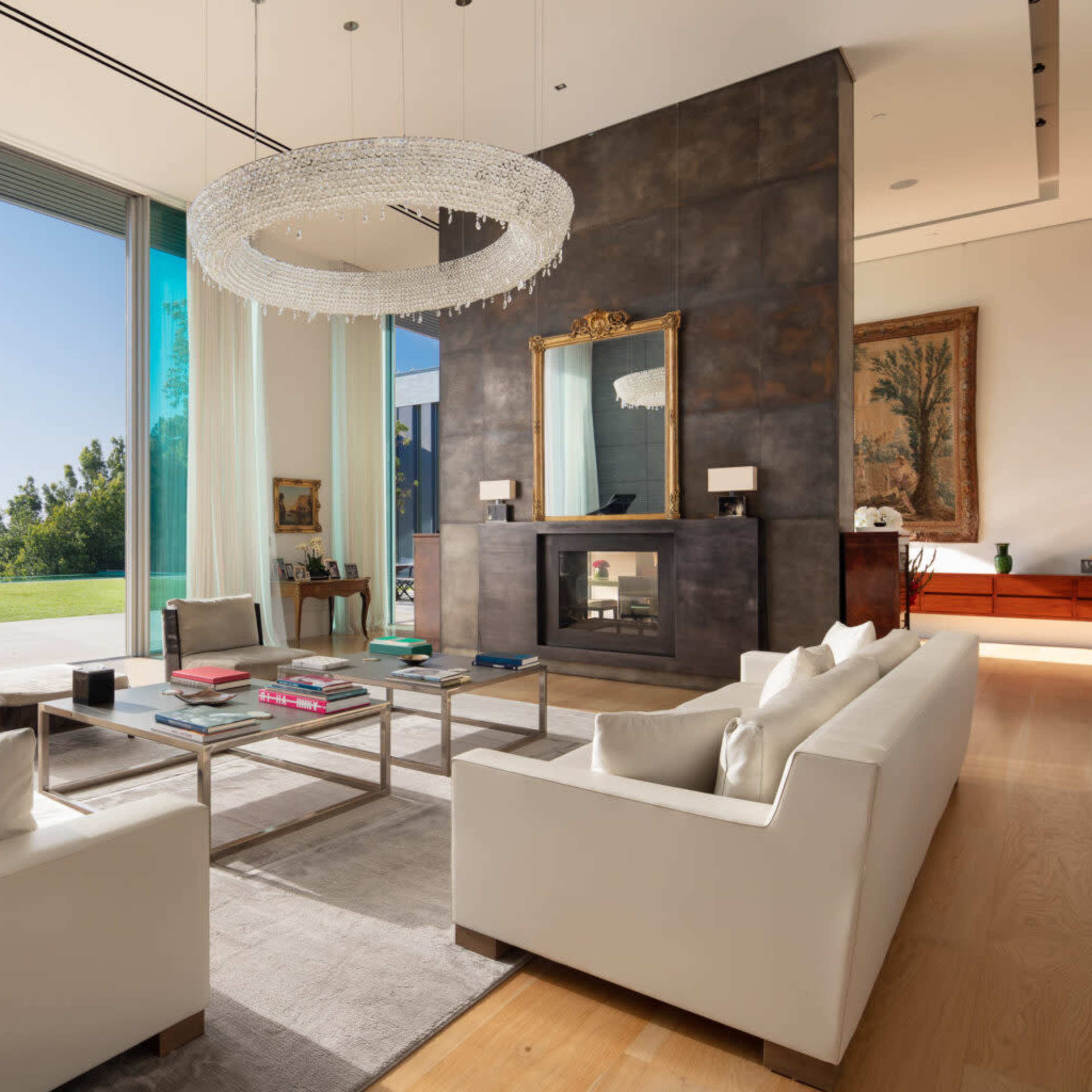A modern living room with large windows, a contemporary chandelier, and white sofas arranged around a coffee table.