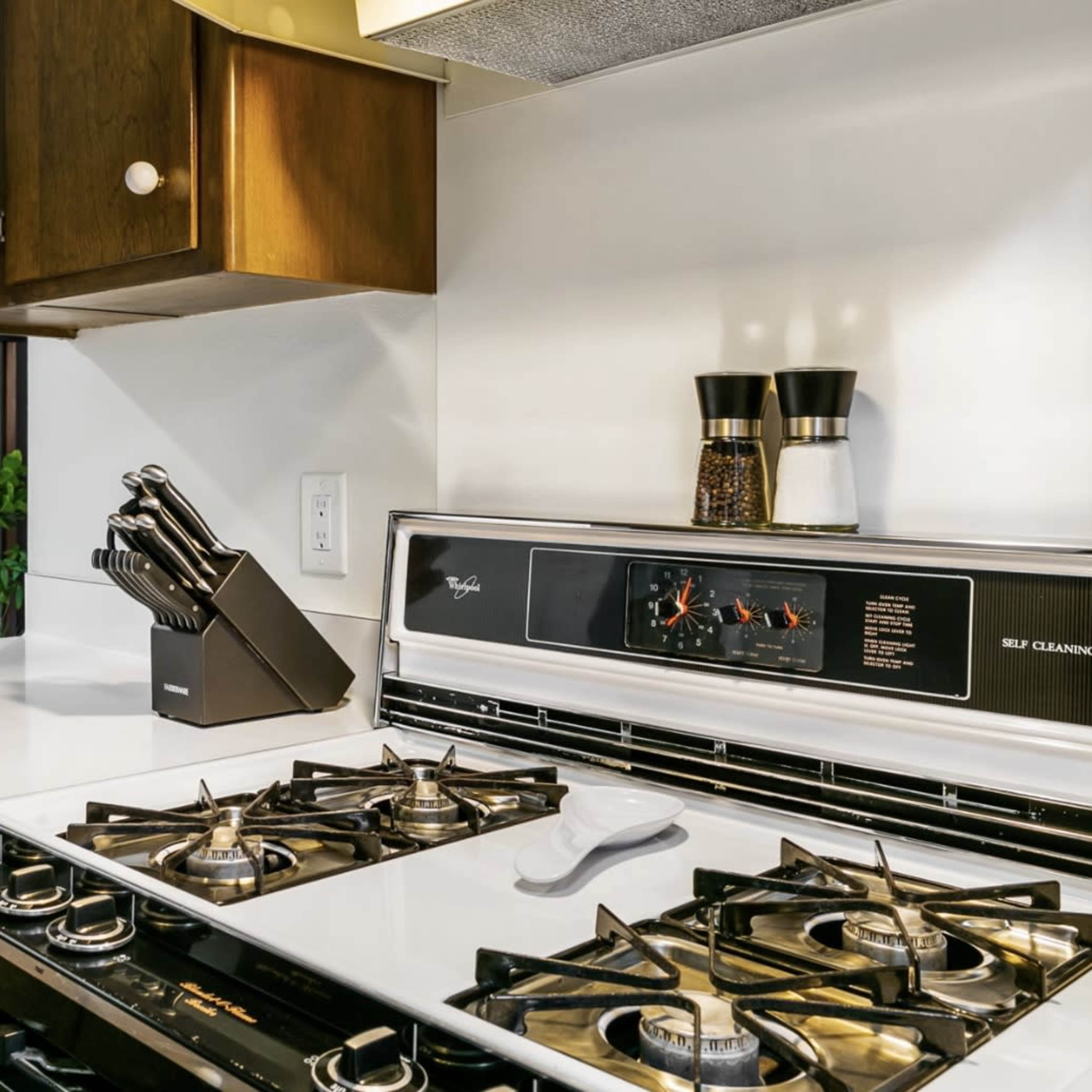 A gas stove with a black knife block sits on a white countertop in a kitchen, adjacent to a window overlooking a patio area.